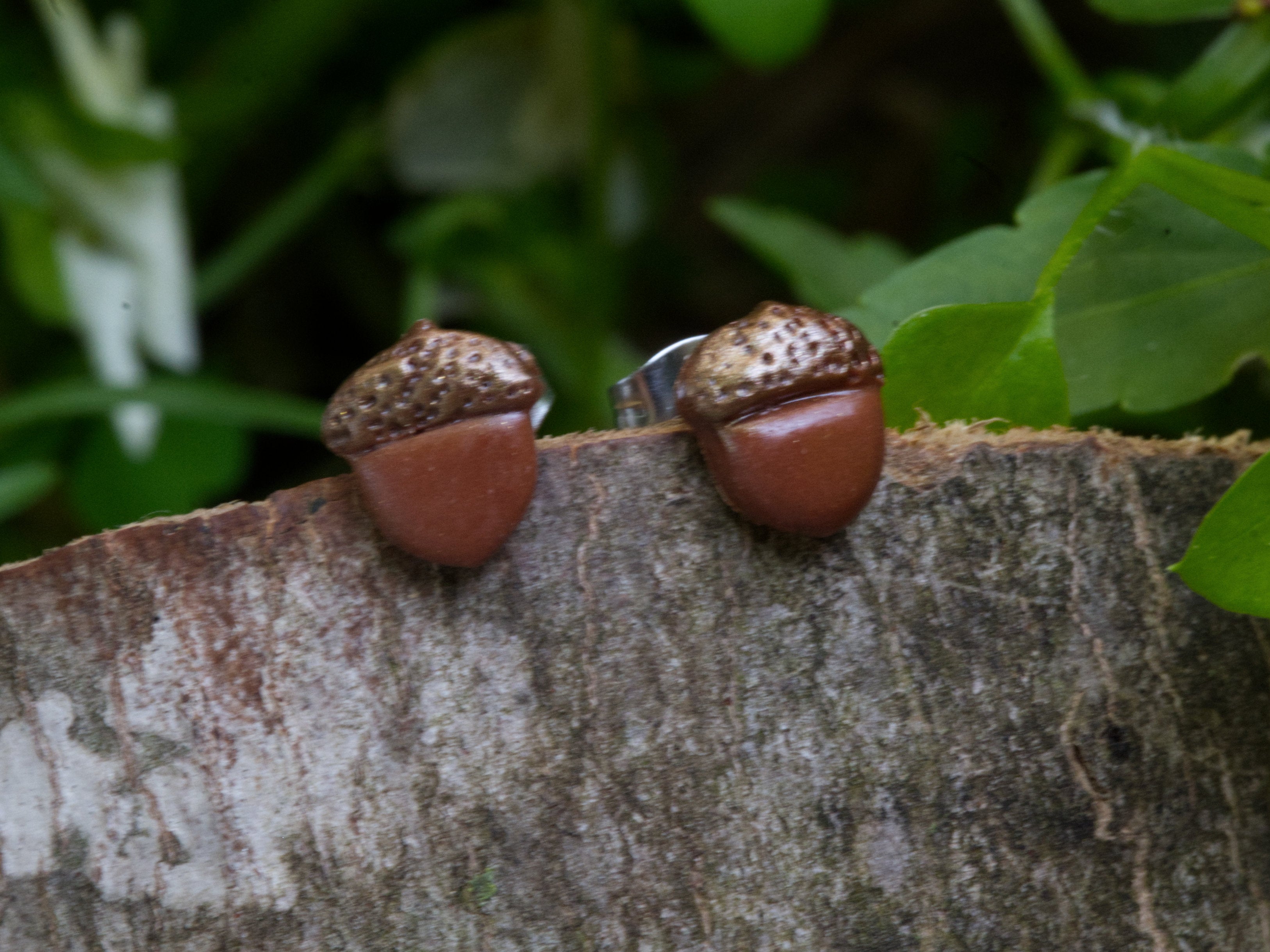 Handmade brown polymer clay acorn stud earrings perched on natural bark surface with green garden foliage background