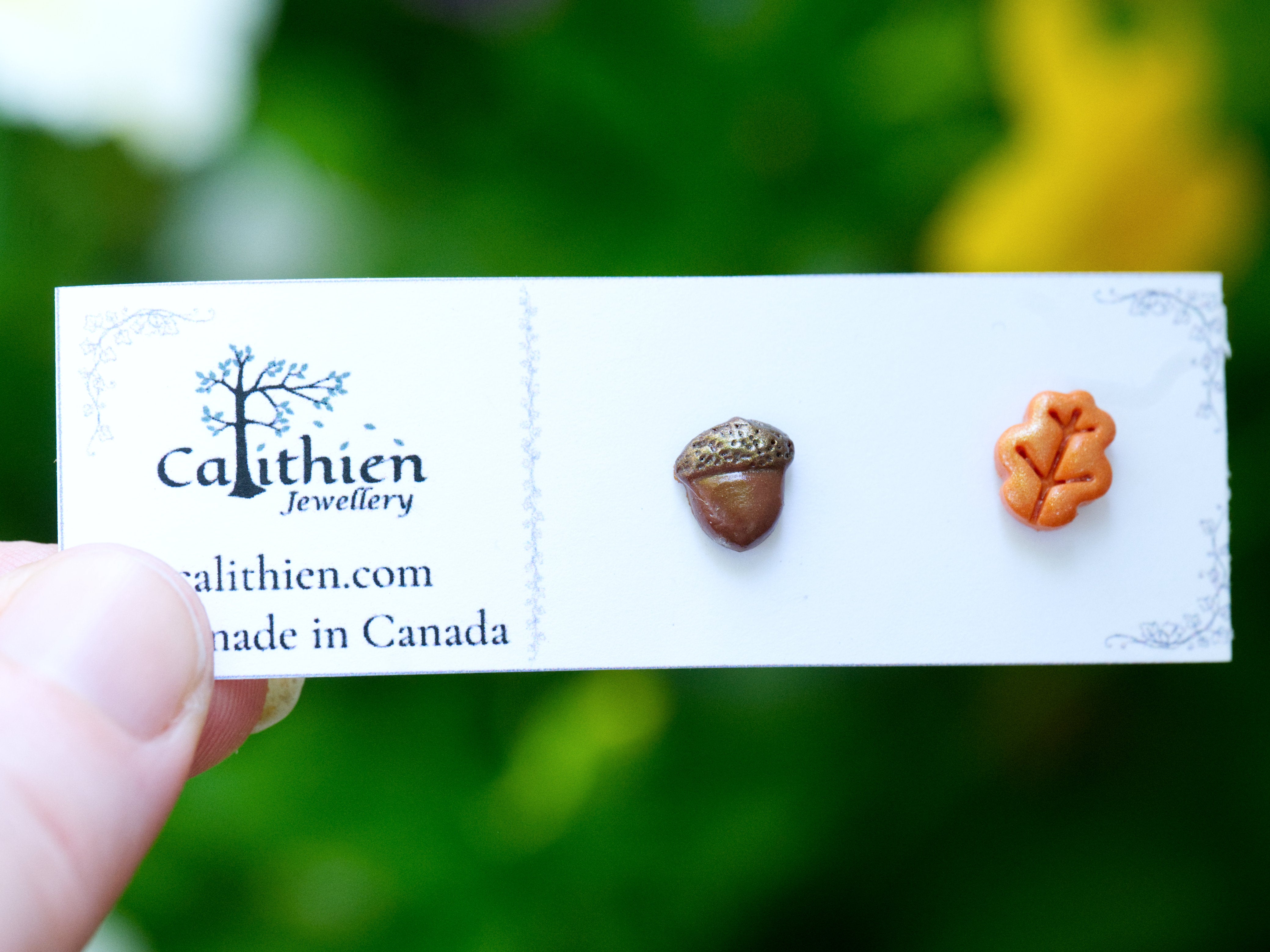 Mismatched oak leaf and acorn stud earrings held in fingers on Calithien Jewellery branded display card reading Handmade in Canada