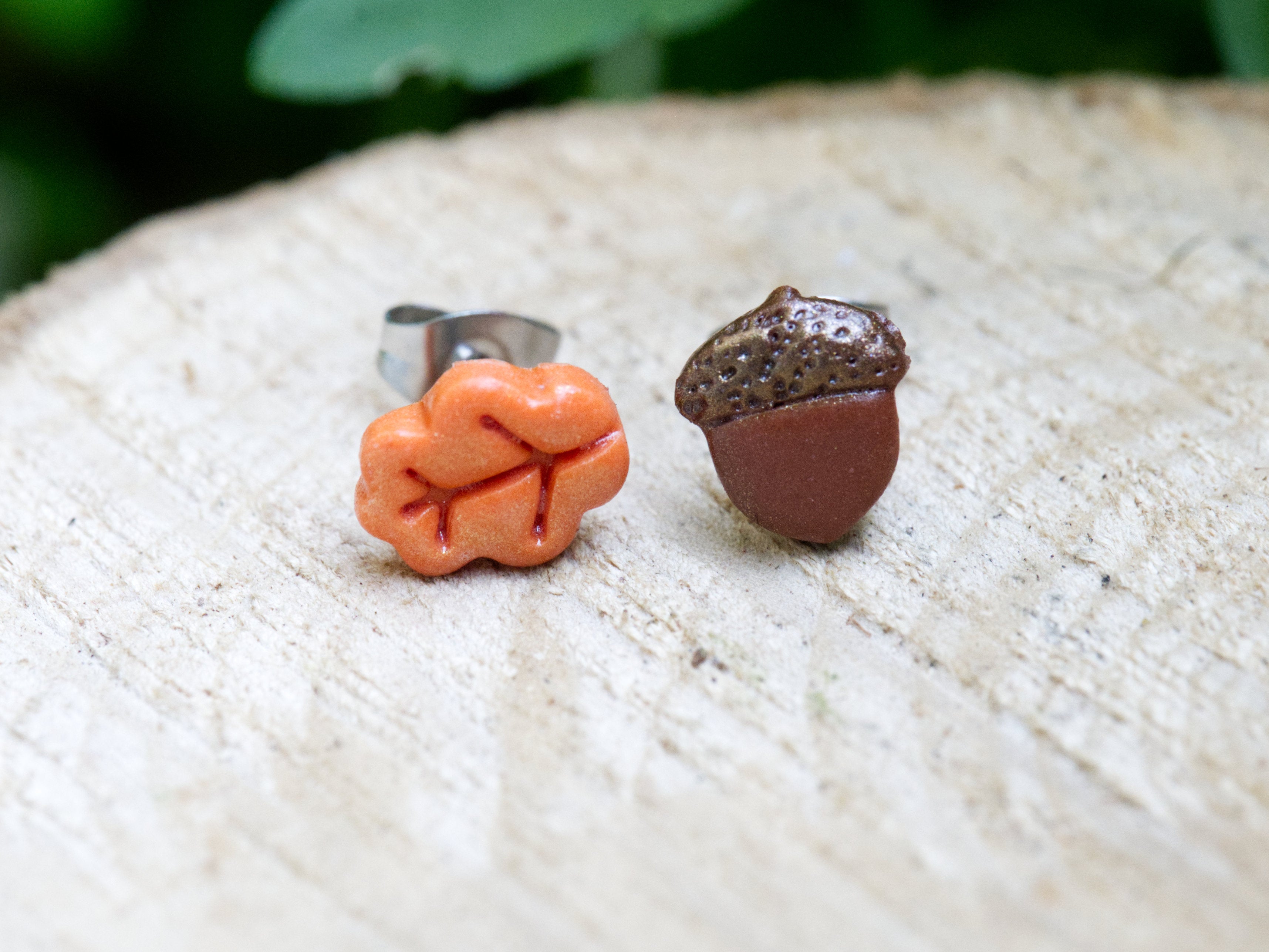 Handmade mismatched orange polymer clay oak leaf and brown acorn stud earrings on natural wooden surface with green garden foliage background