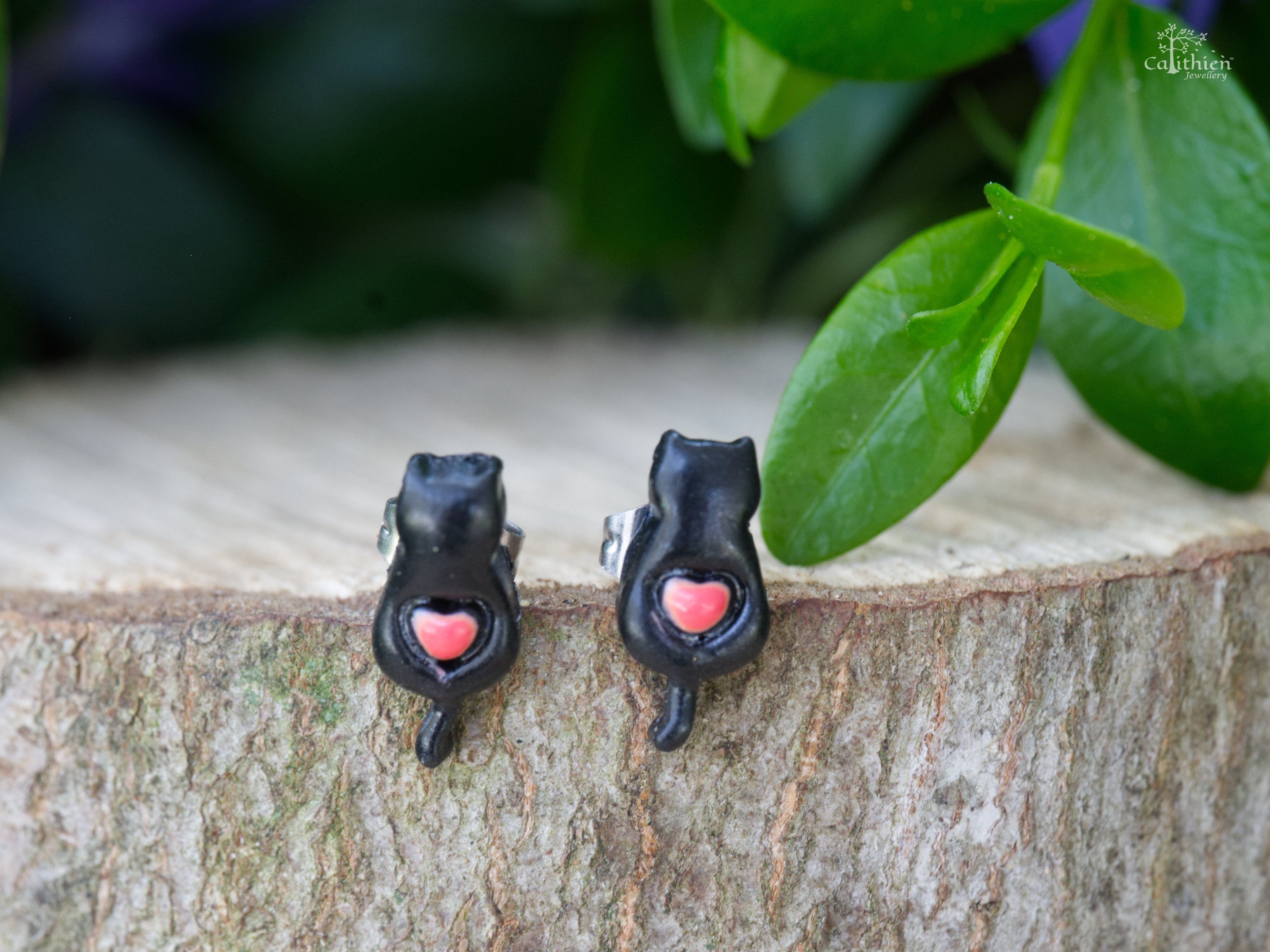 Handmade black polymer clay cat stud earrings with pink heart detail perched on edge of natural wood slice with green leaf background