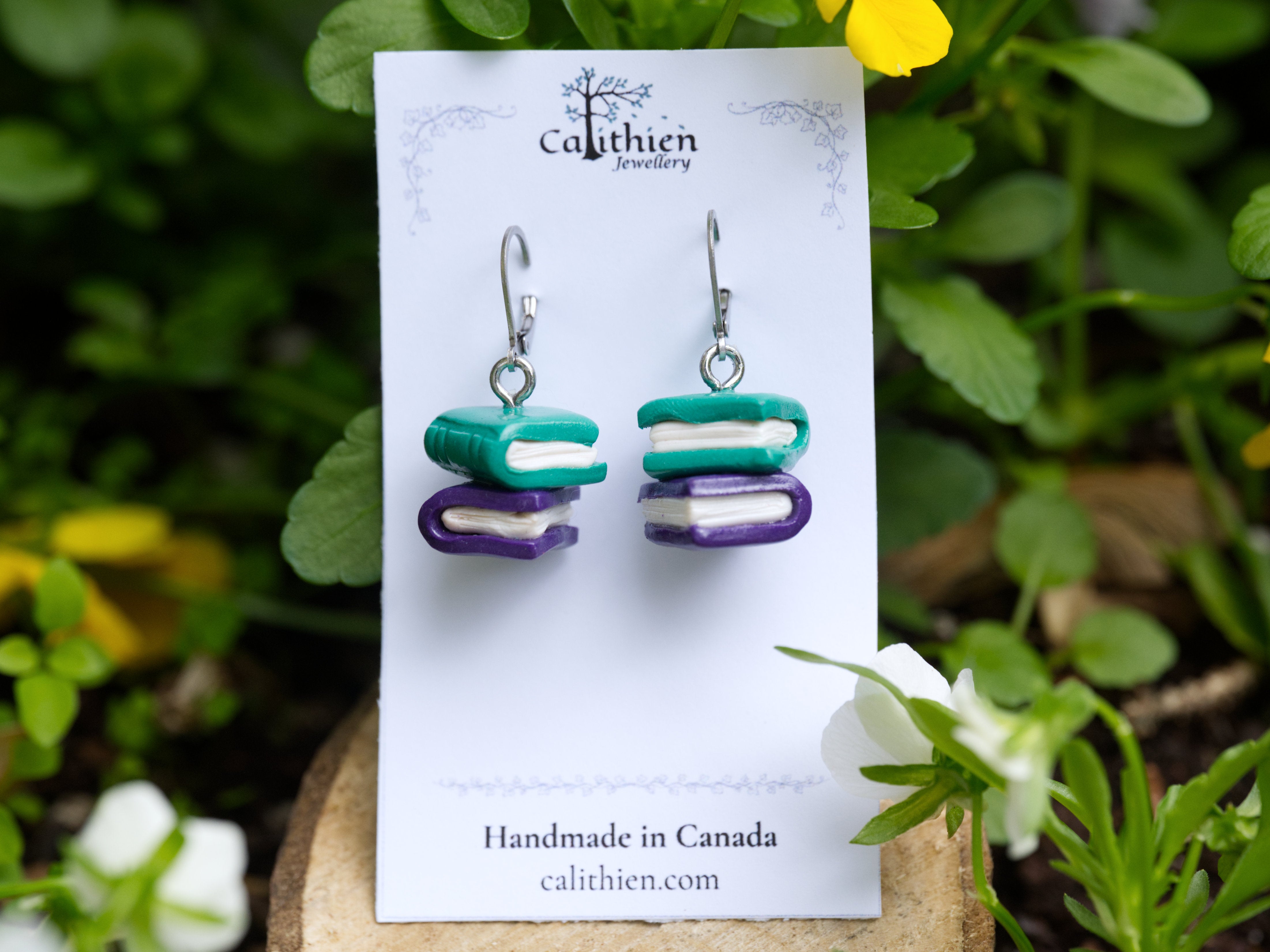 Teal and purple handmade polymer clay book stack earrings on silver leverback hooks displayed on Calithien Jewellery branded card with garden flowers in background