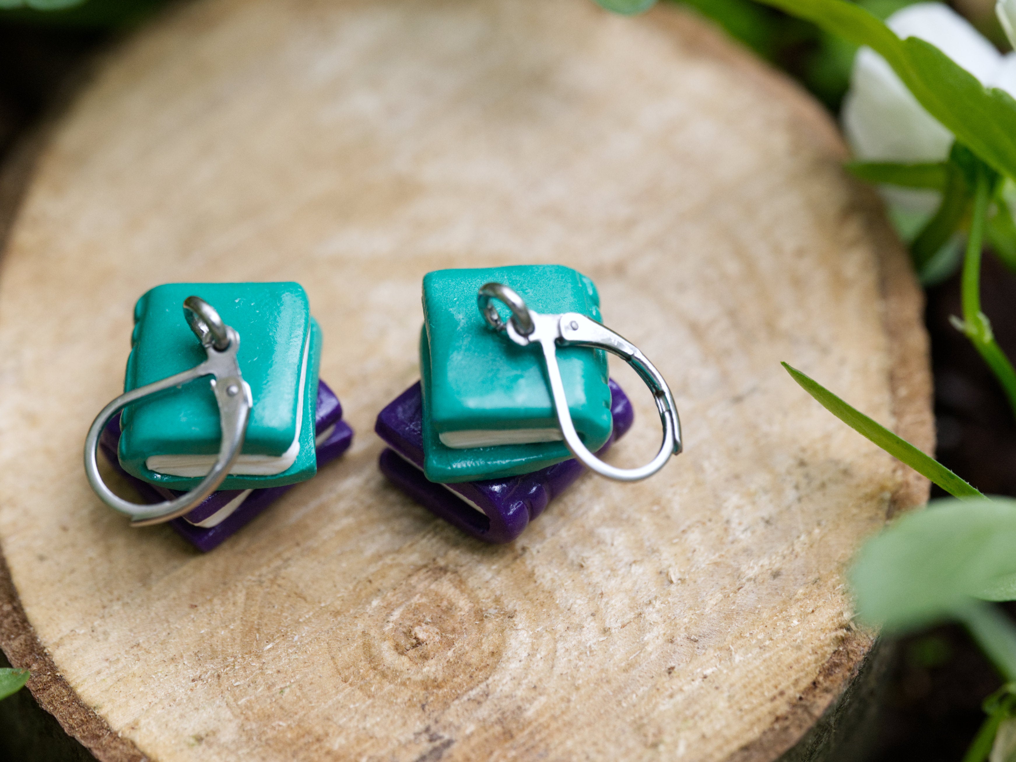 Teal and purple book stack earrings resting on natural wood slice surrounded by green leaves and white flowers in soft natural daylight