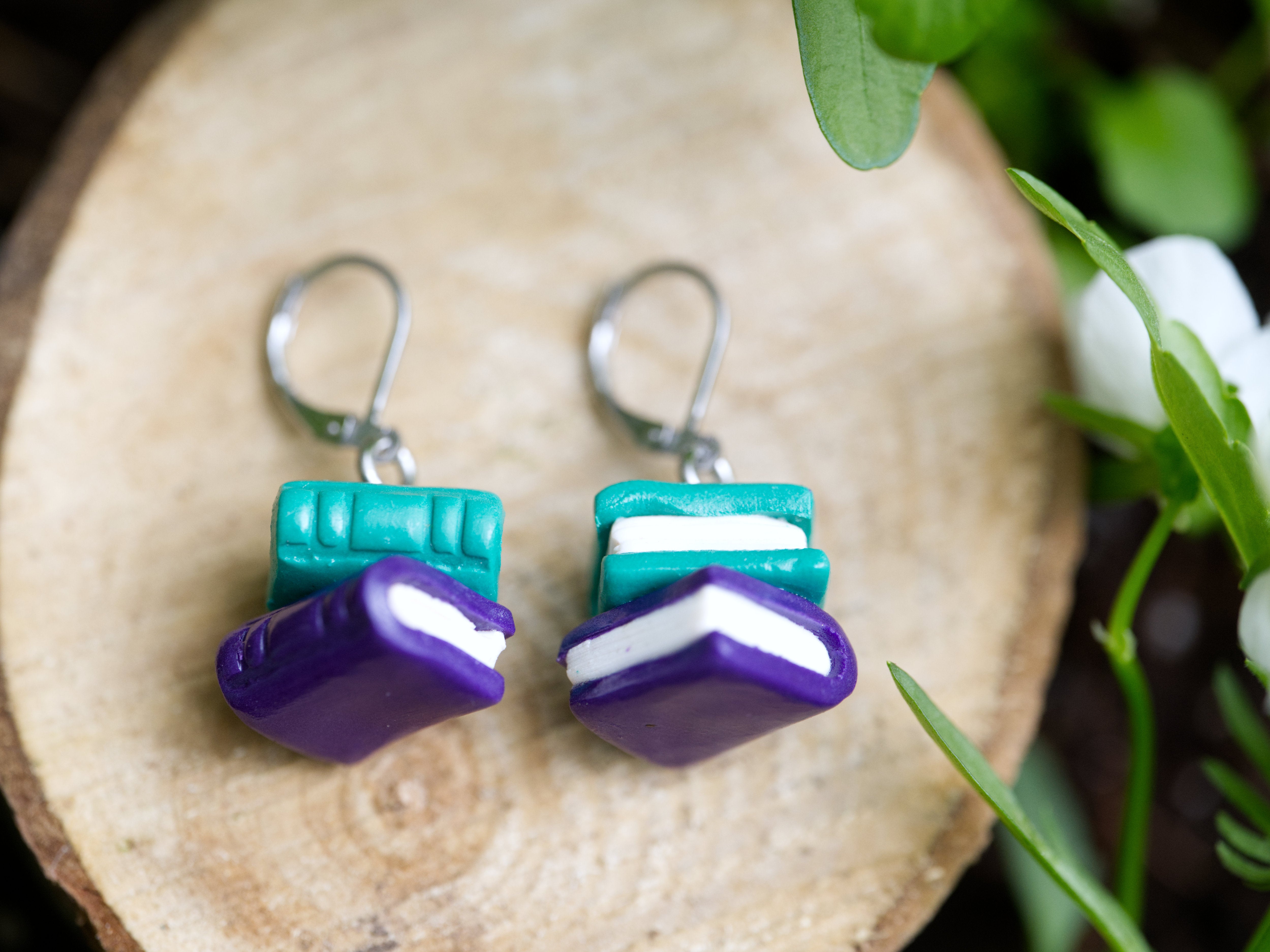 Close-up of handmade book stack earrings showing sculpted page detail, teal and purple polymer clay, and silver leverback hooks