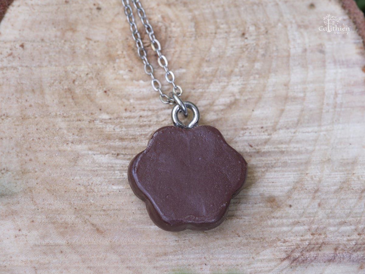 Back view of the handmade brown polymer clay paw print necklace pendant displayed on a natural wooden surface.