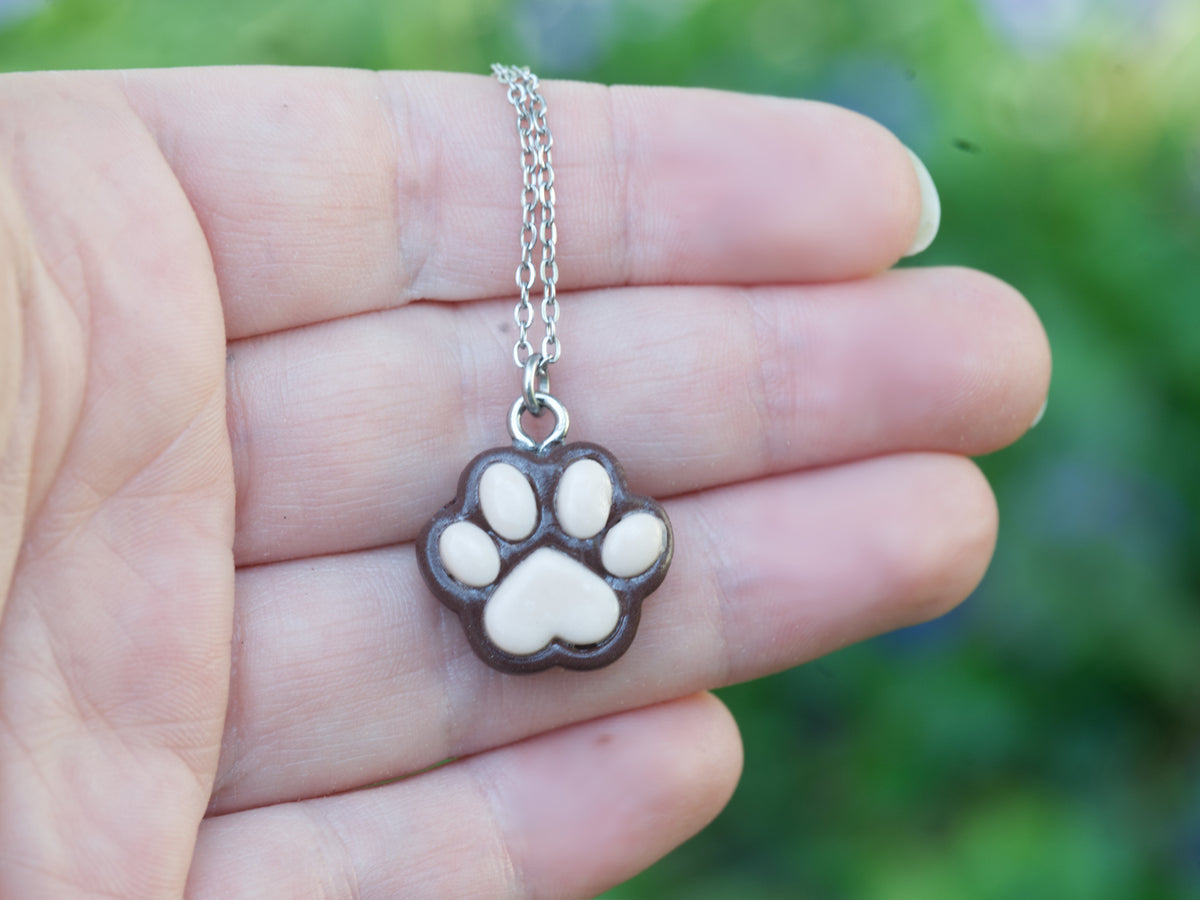 Brown paw print necklace with pink pads resting on a hand against a softly blurred greenery background.