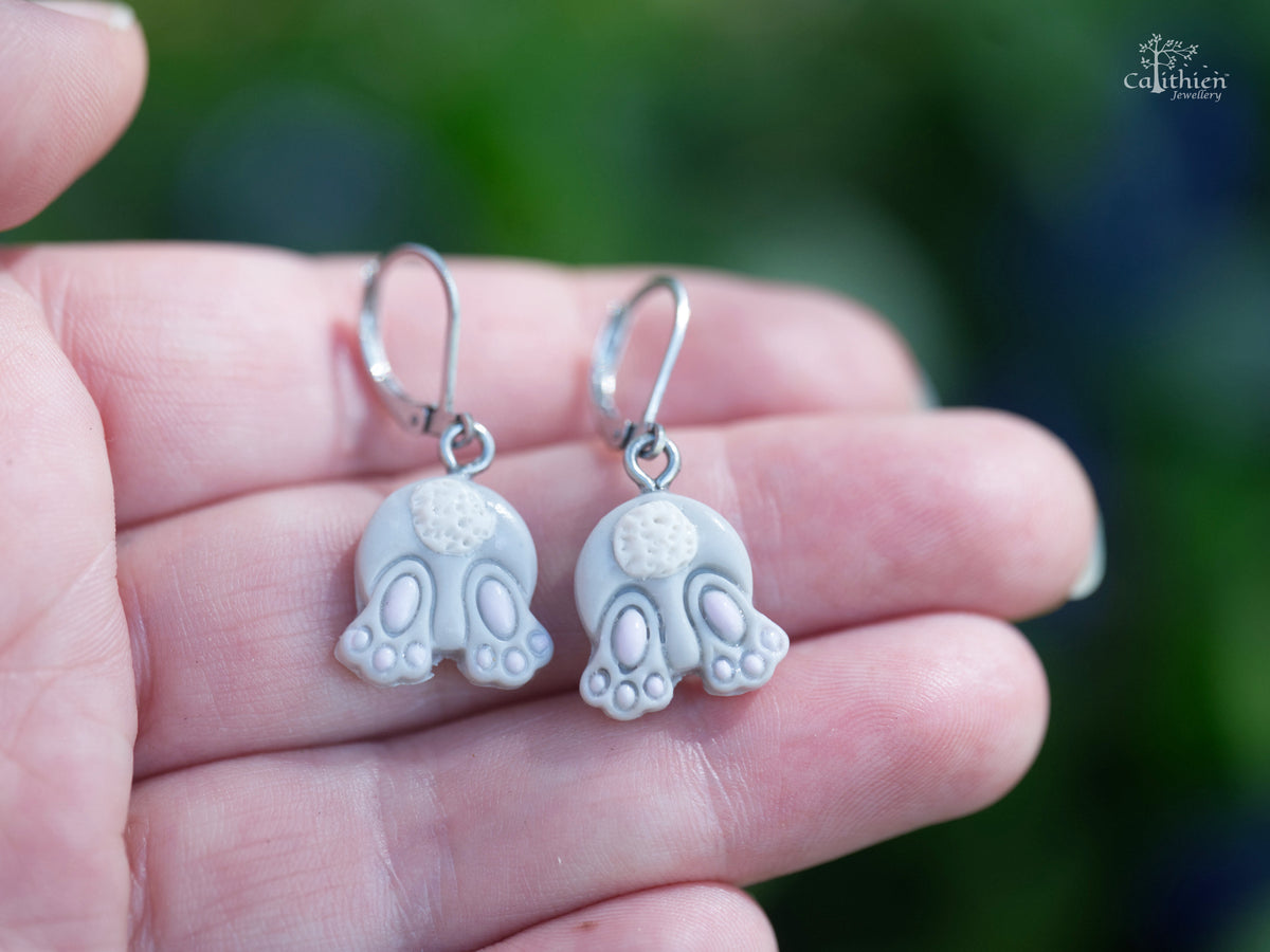 Grey bunny bum dangle earrings with charms shown against a hand to demonstrate size