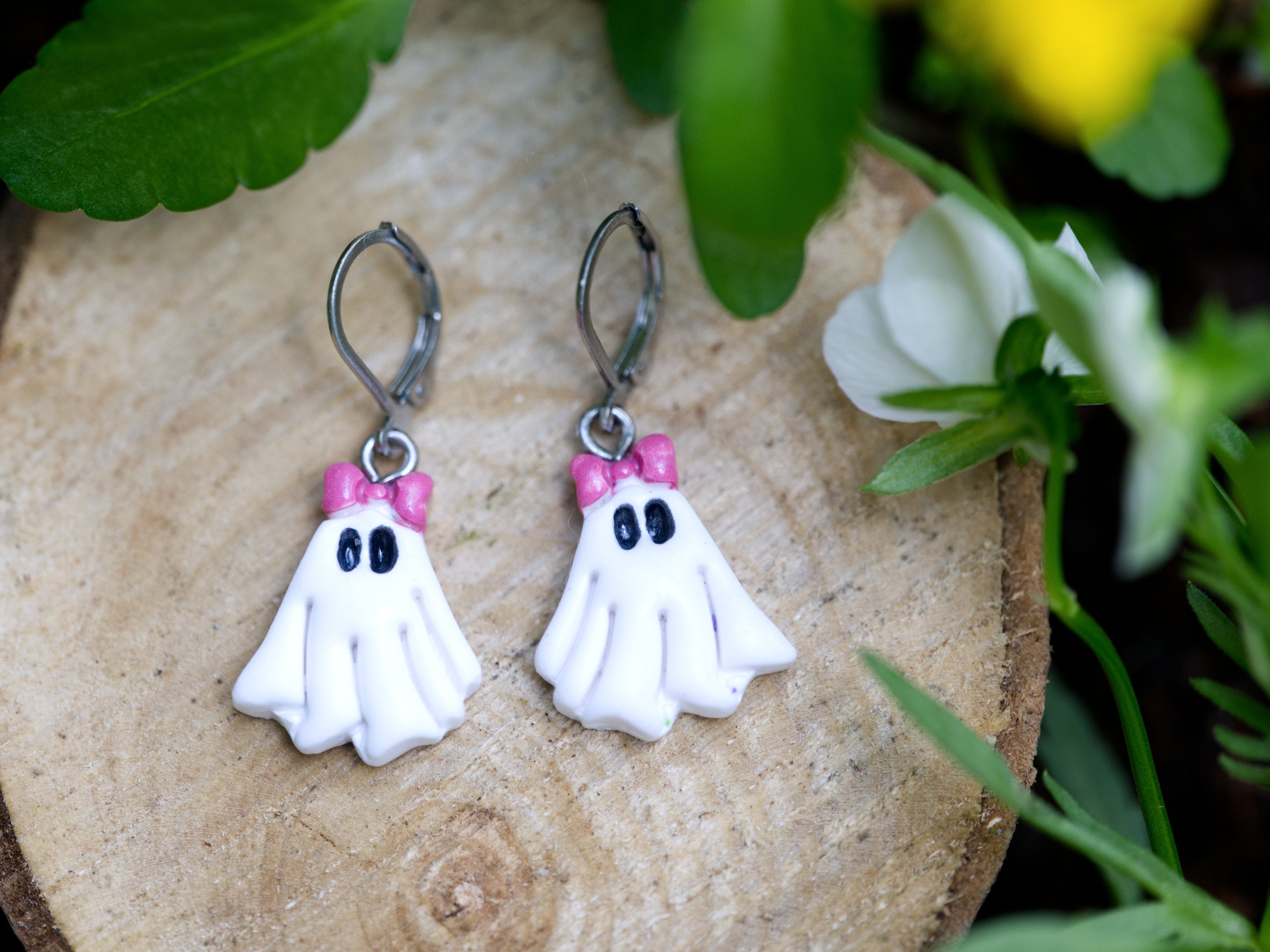 Handmade white polymer clay coquette ghost leverback earrings with pink bow detail on natural wood slice with garden flowers background