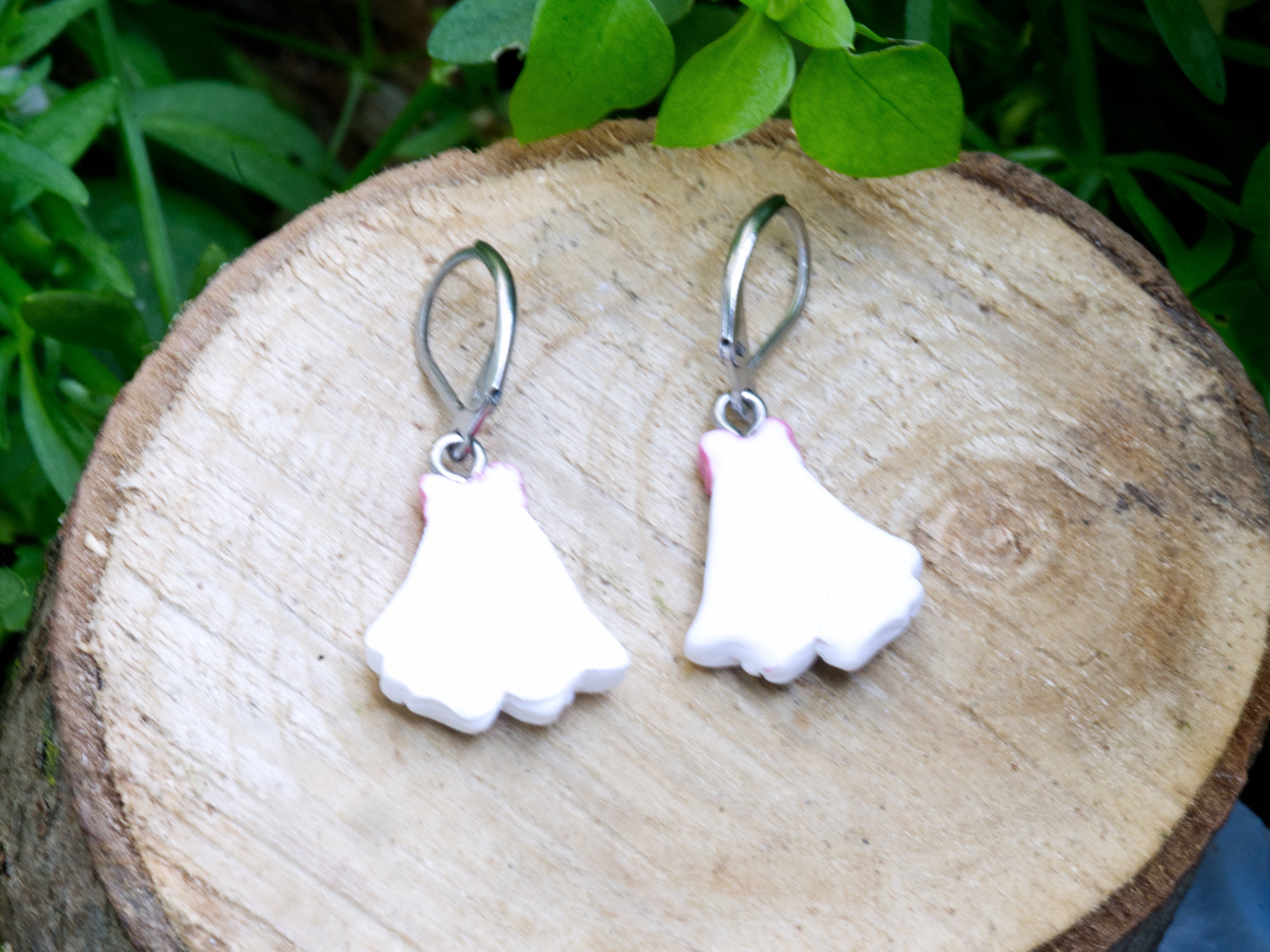Back of handmade white polymer clay coquette ghost leverback earrings with pink bow detail on natural wood slice with garden flowers background