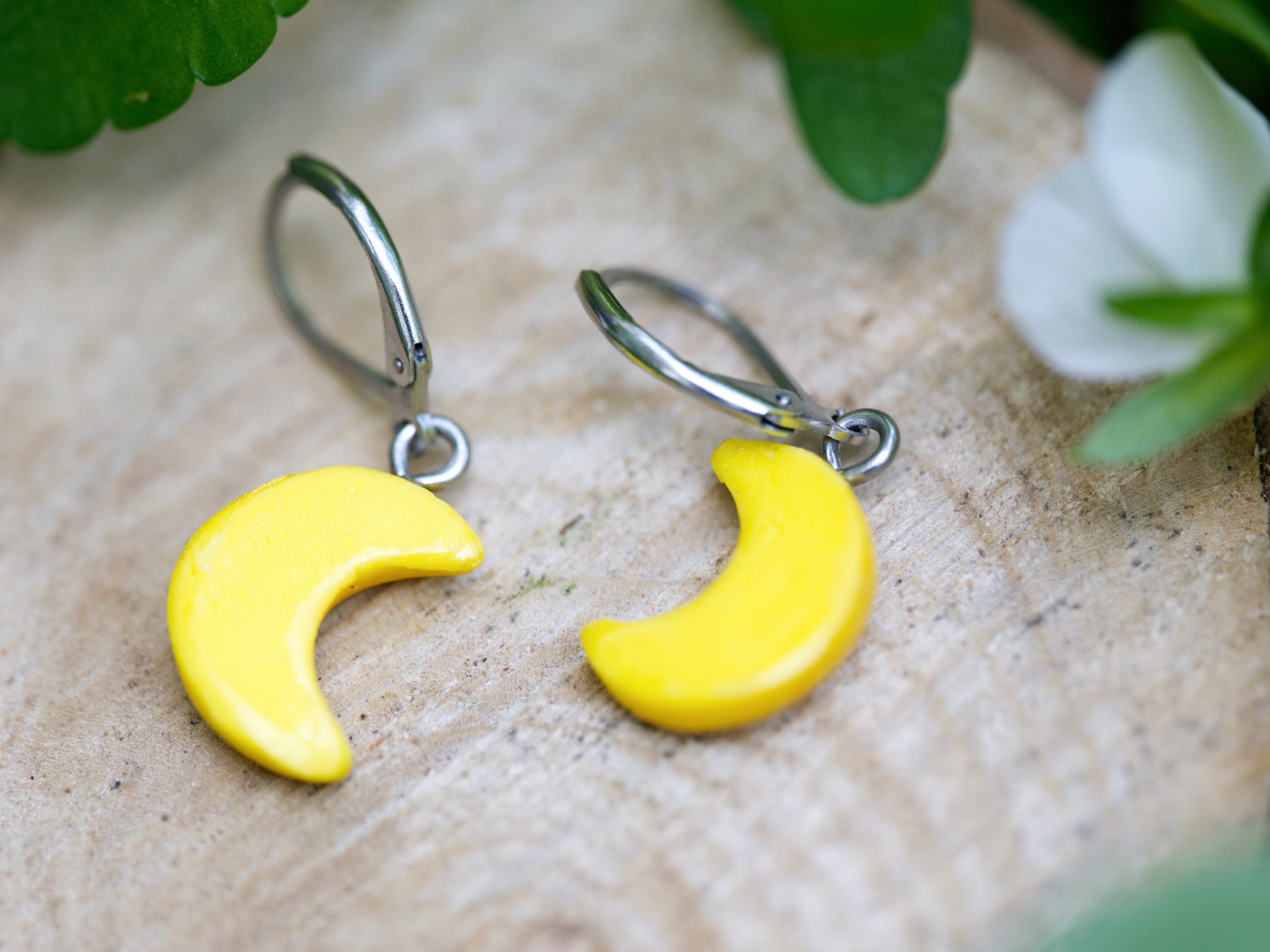 Backs of gold crescent moon earrings on wood slice with soft green foliage and white flower bokeh background