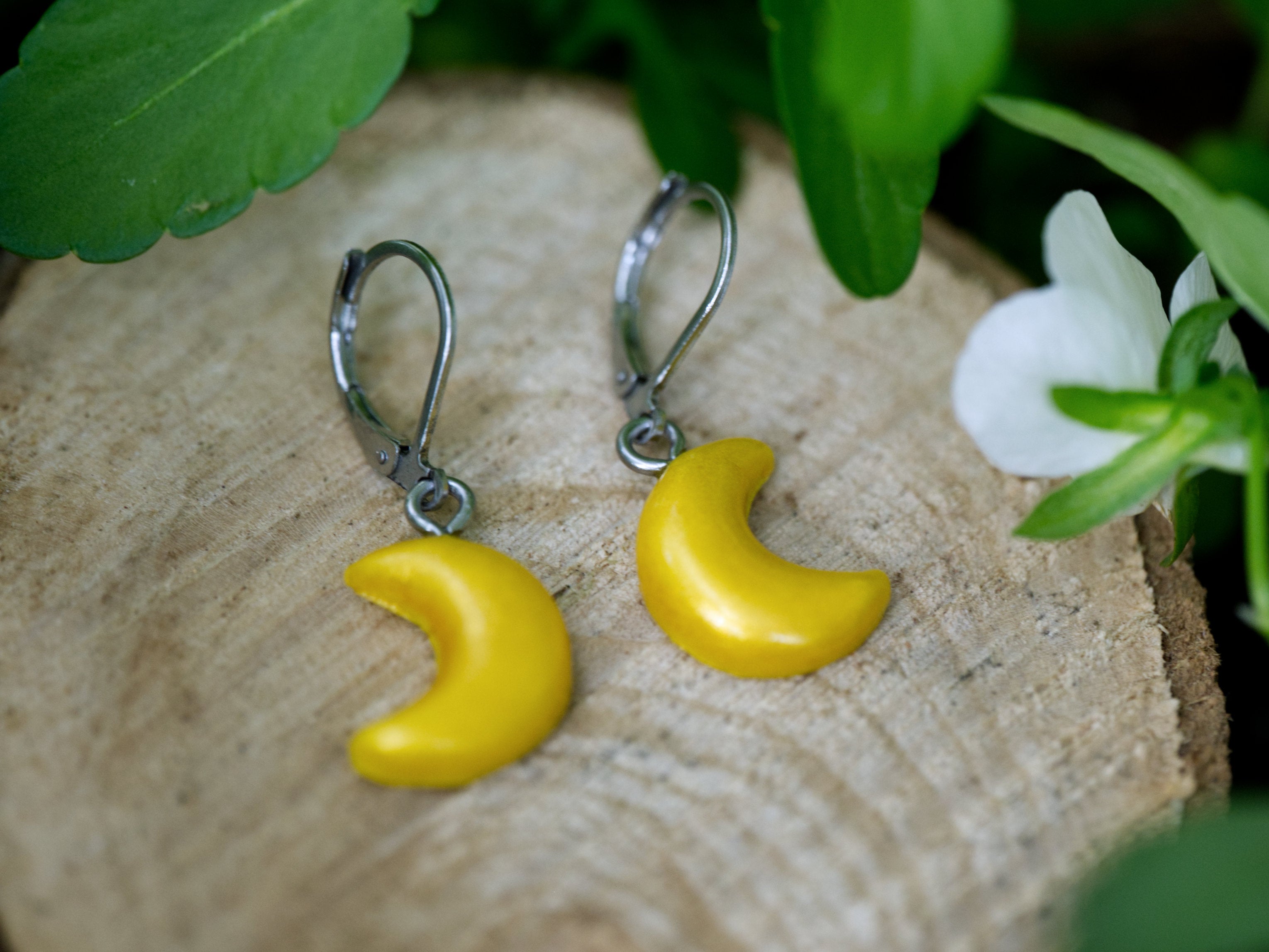 Pair of gold crescent moon earrings on wood slice with soft green foliage and white flower bokeh background