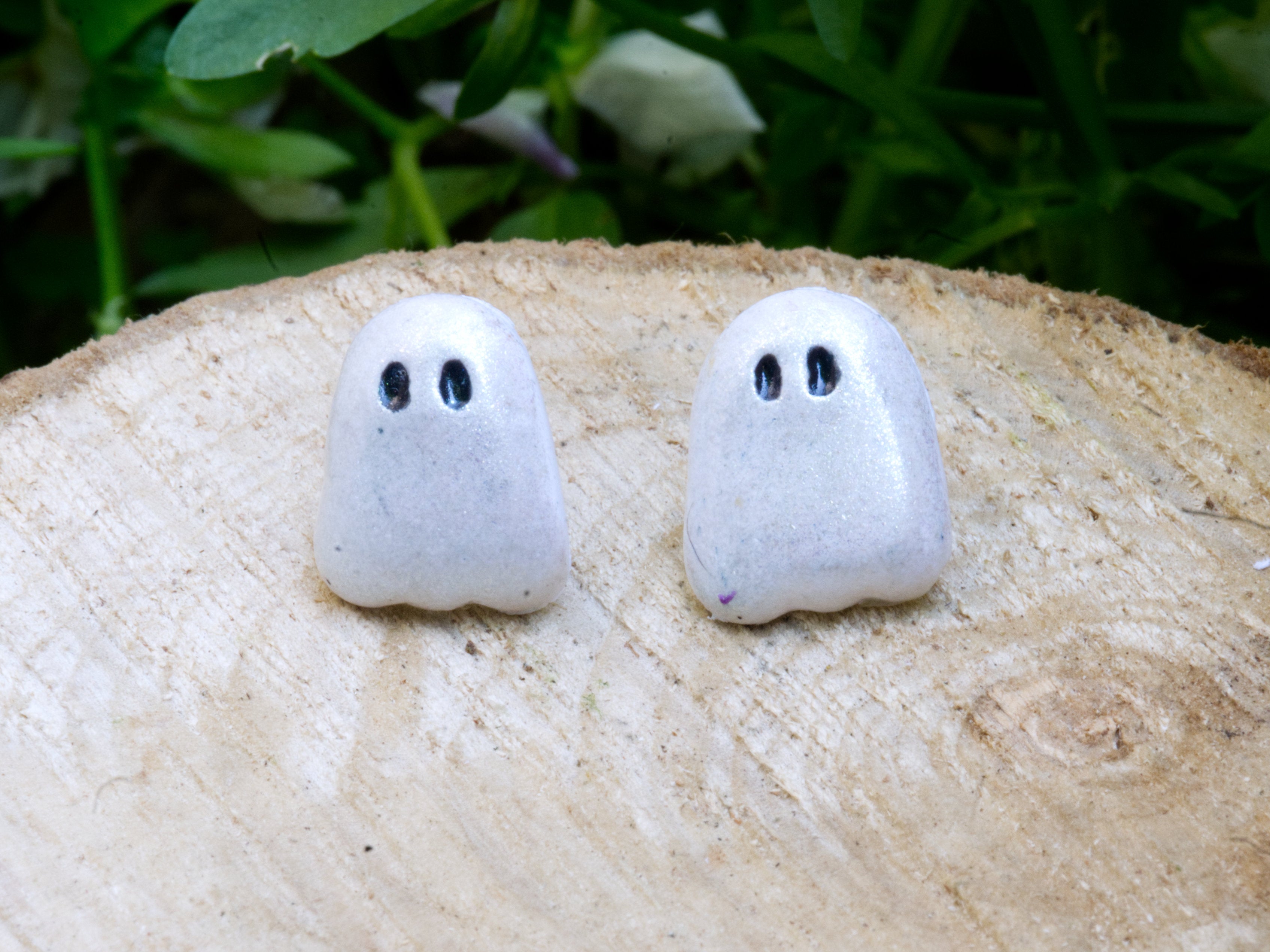 Pair of ghost stud earrings on wood slice surface with soft green bokeh background in natural daylight