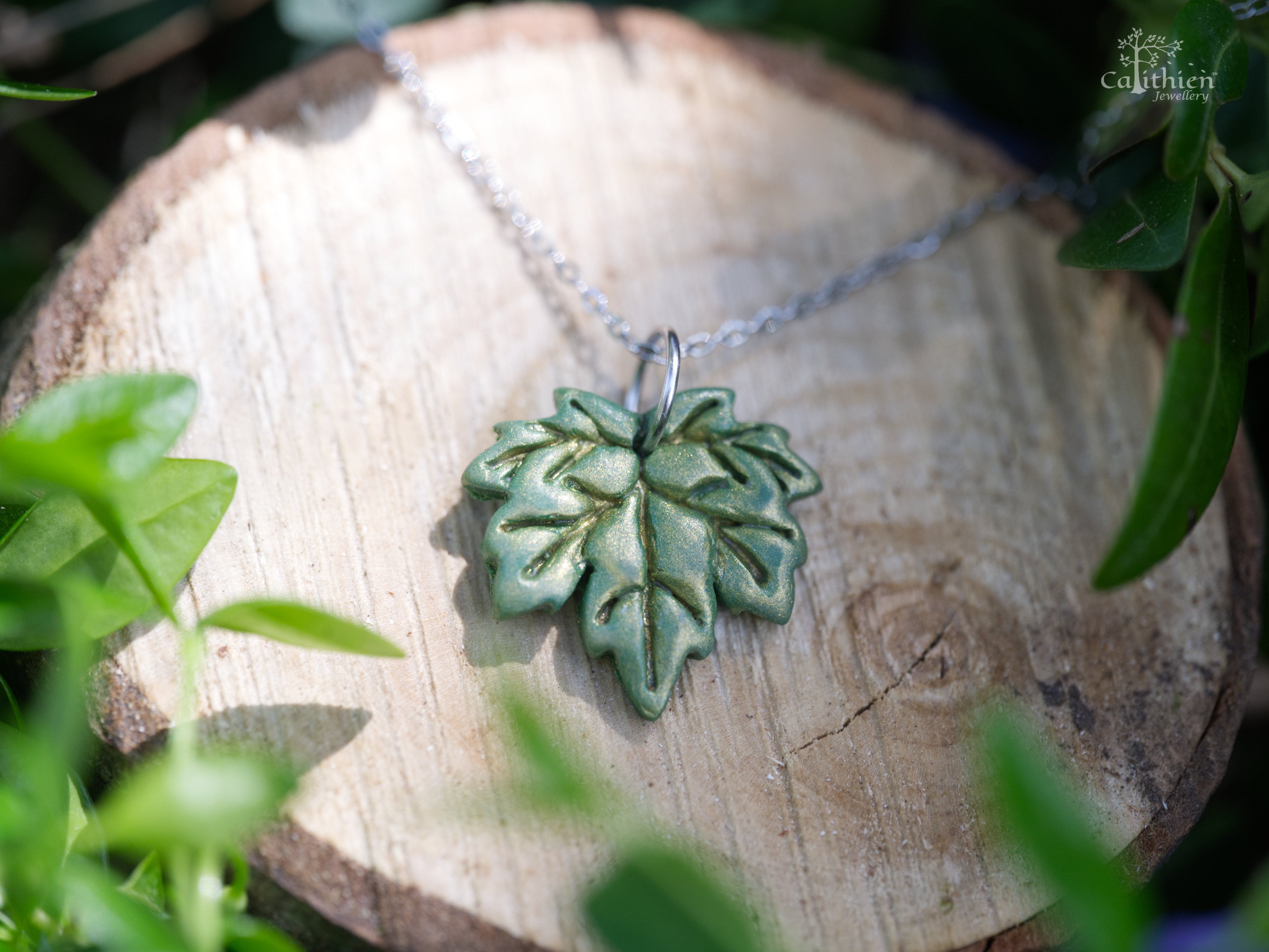 Handmade green shimmer polymer clay maple leaf pendant necklace on 304 stainless steel chain resting on natural wood slice with green foliage