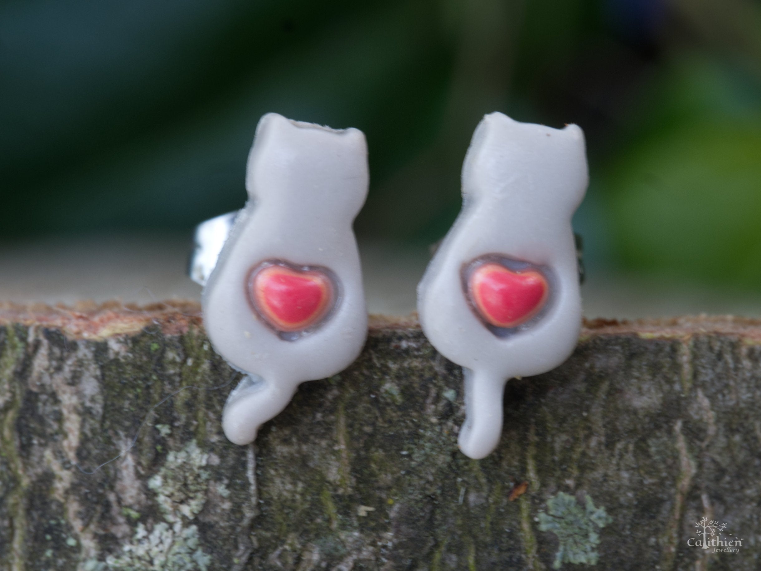 Handmade grey polymer clay cat stud earrings with pink heart detail on natural wood surface with green garden backdrop