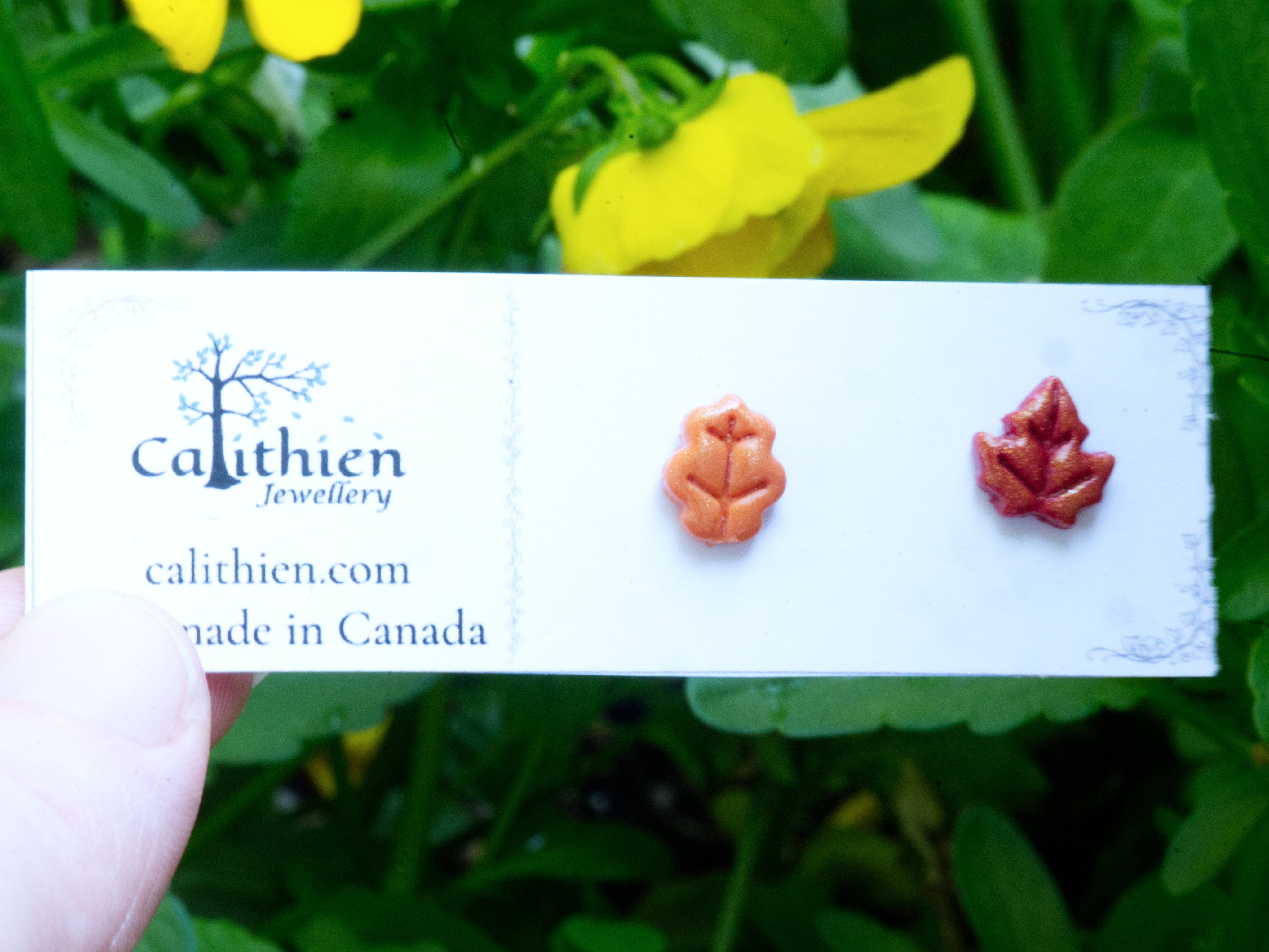 Mismatched oak and maple leaf stud earrings behing held on a Calithien Jewellery branded display card reading Handmade in Canada