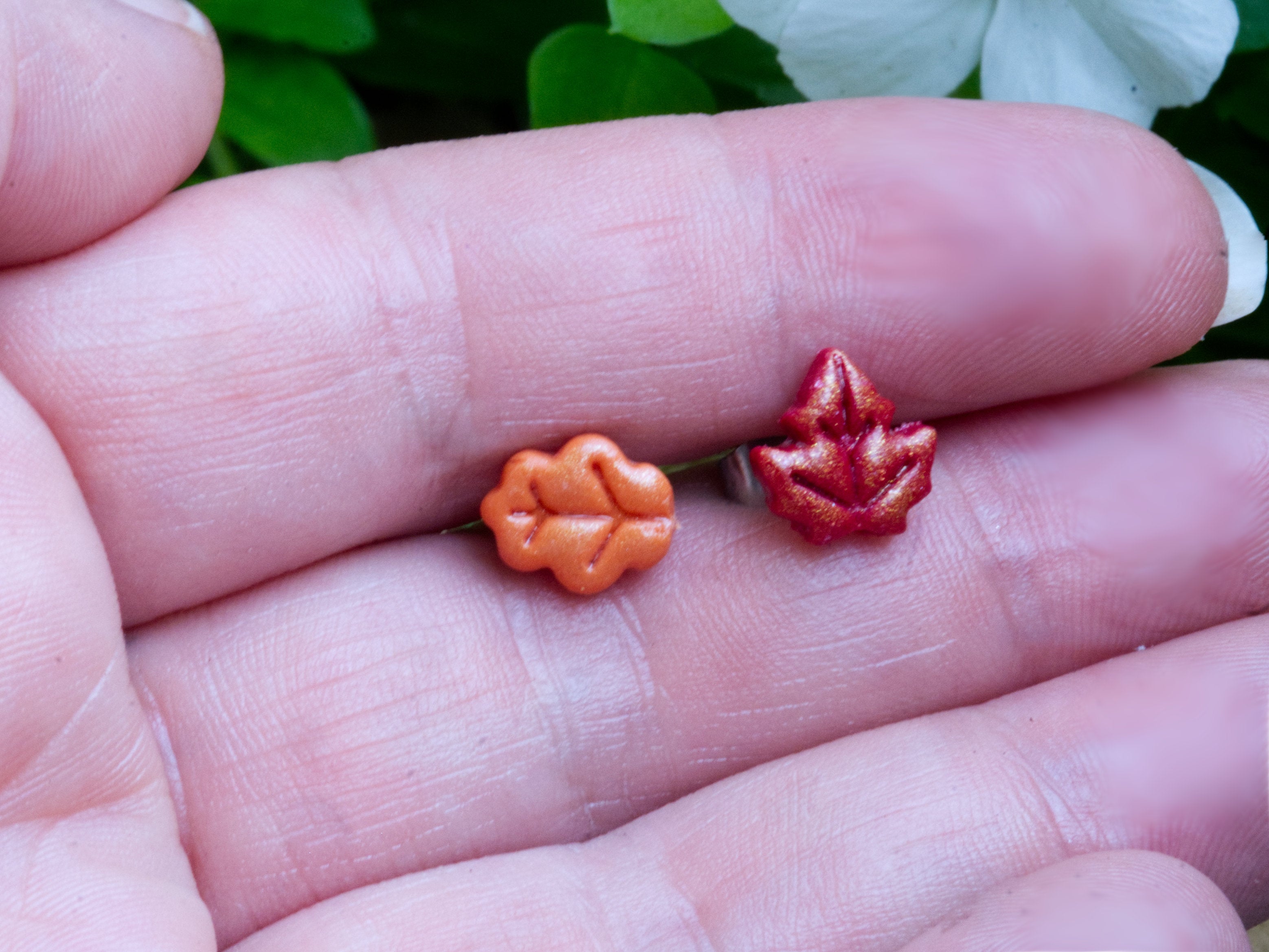 Orange and red polymer clay leaf stud earrings at true size approximately 1 centimetre each shown on a hand