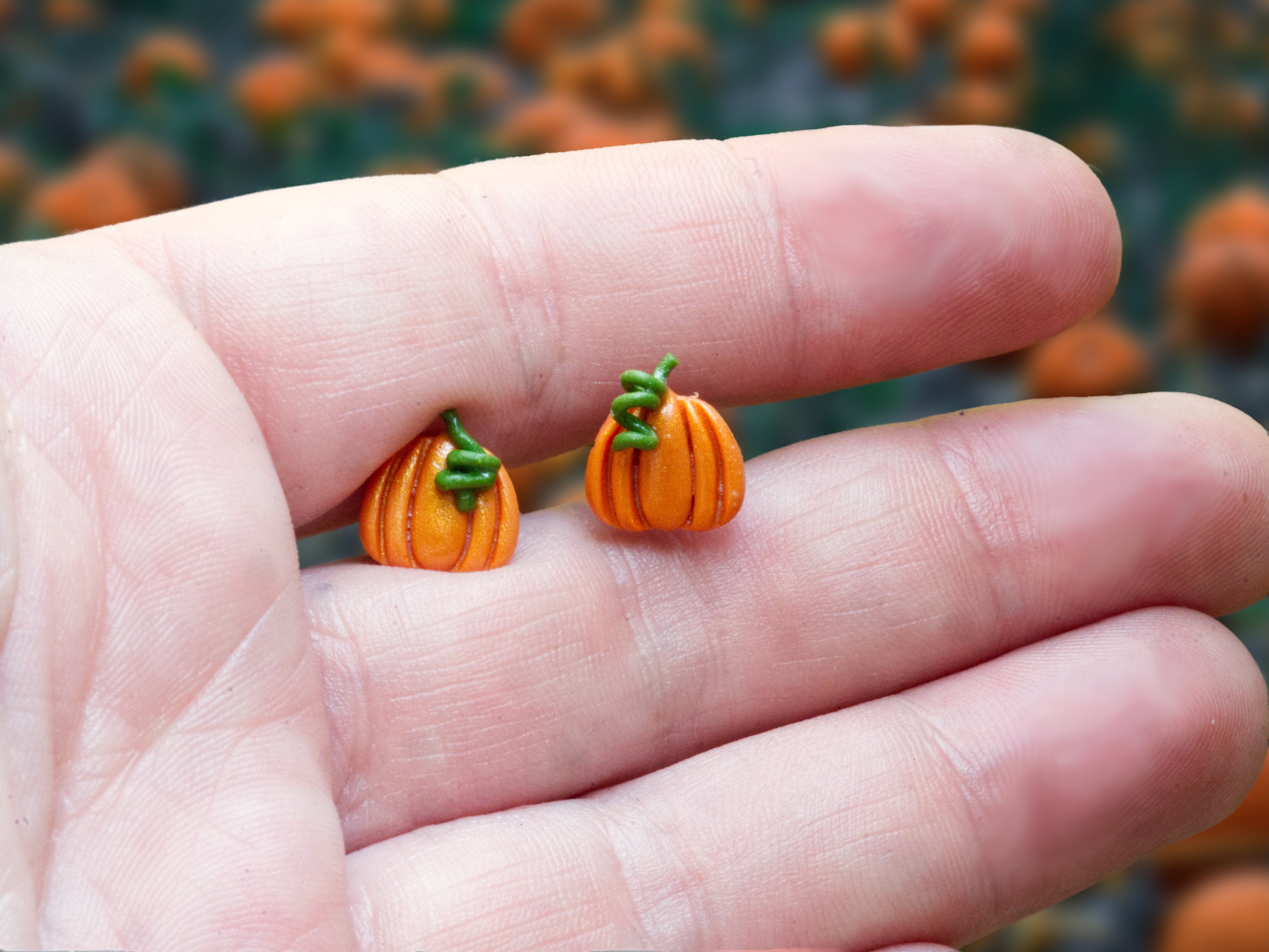 Small orange polymer clay pumpkin stud earrings held in a hand to show their approximately 1.5 cm size and delicate, lightweight design.
