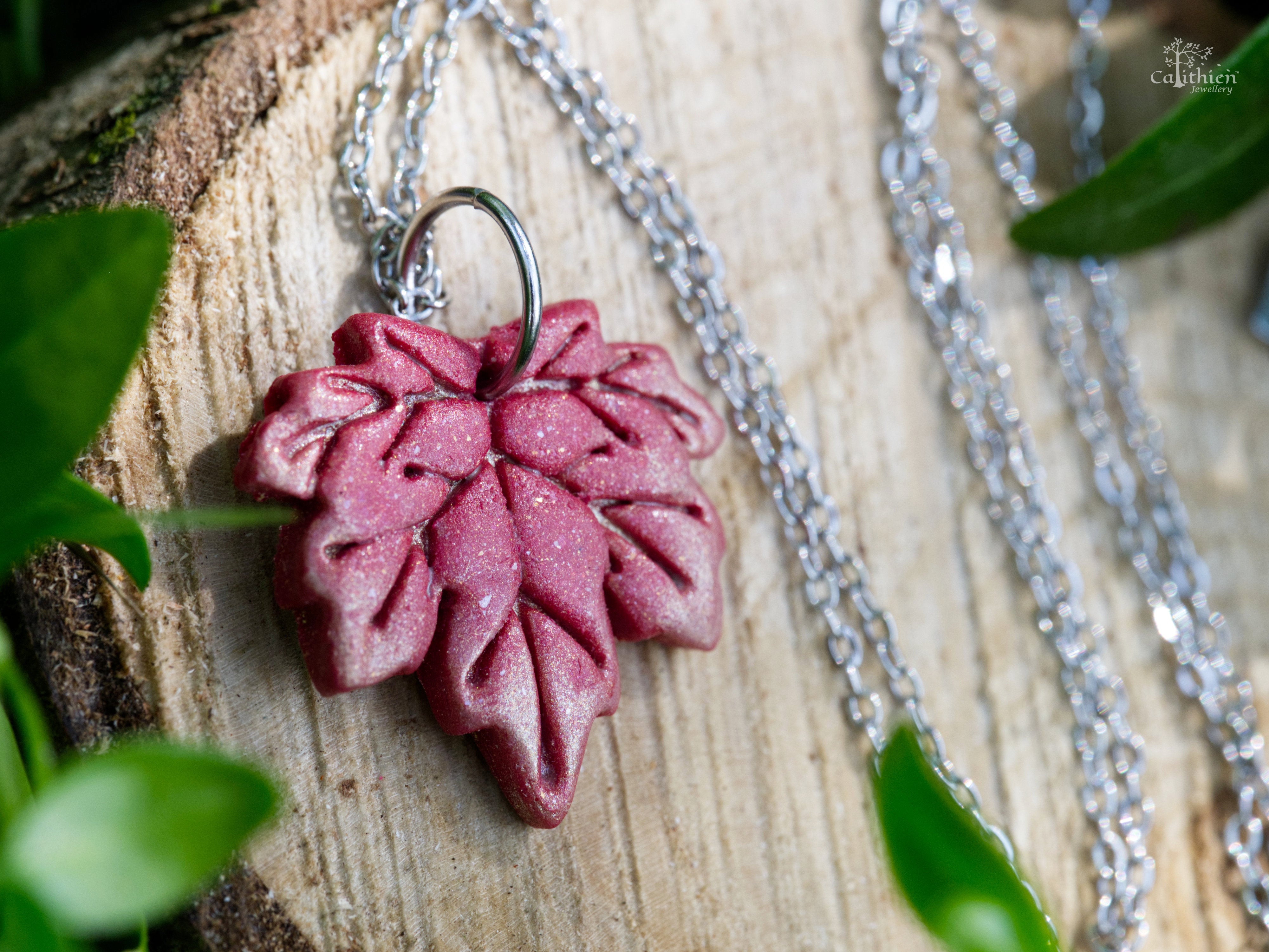 Handmade red and gold shimmer polymer clay maple leaf pendant necklace on 304 stainless steel chain resting on natural wood slice with green foliage