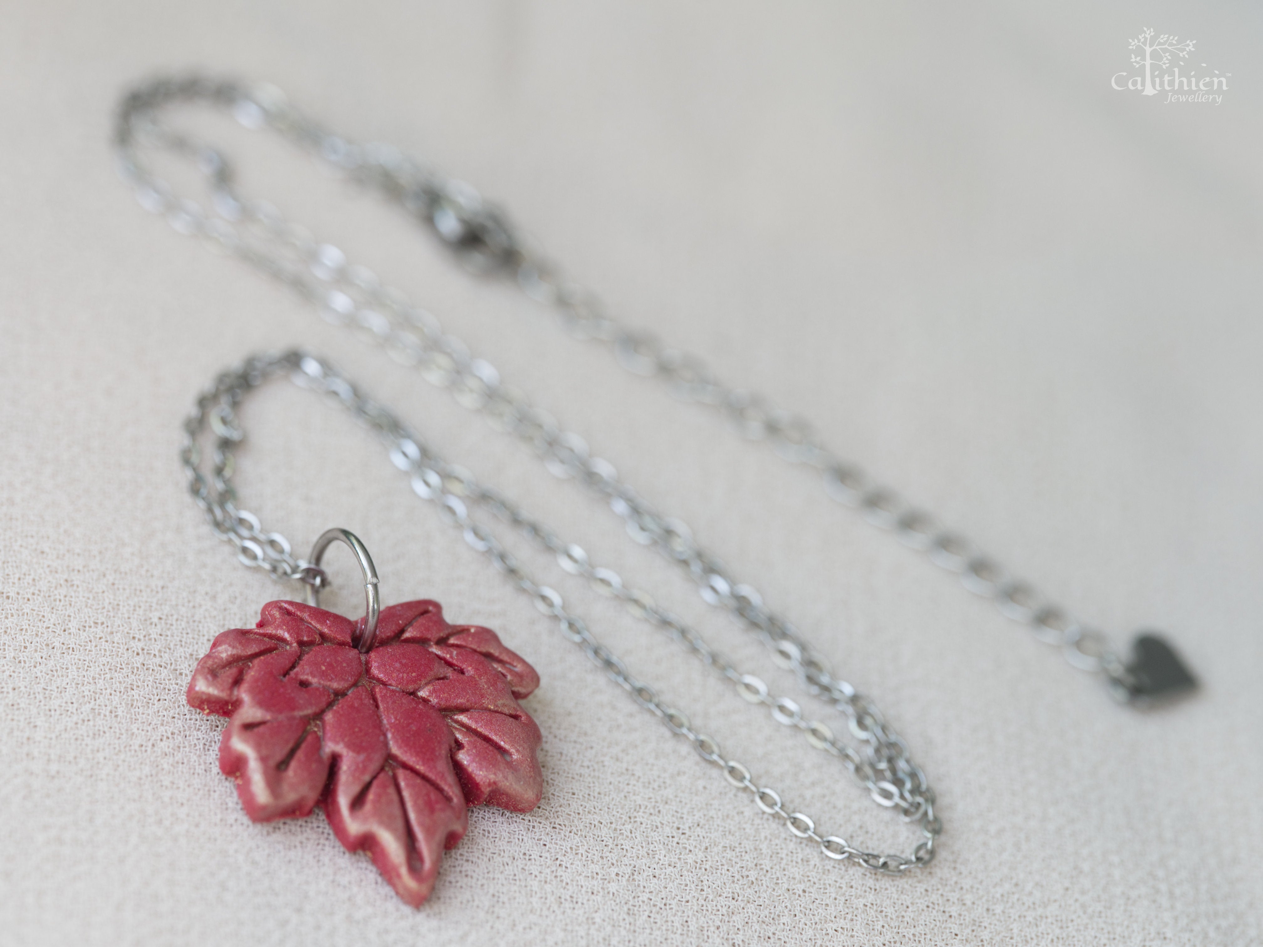 Handmade red and gold shimmer polymer clay maple leaf pendant necklace on 304 stainless steel chain resting on white fabric