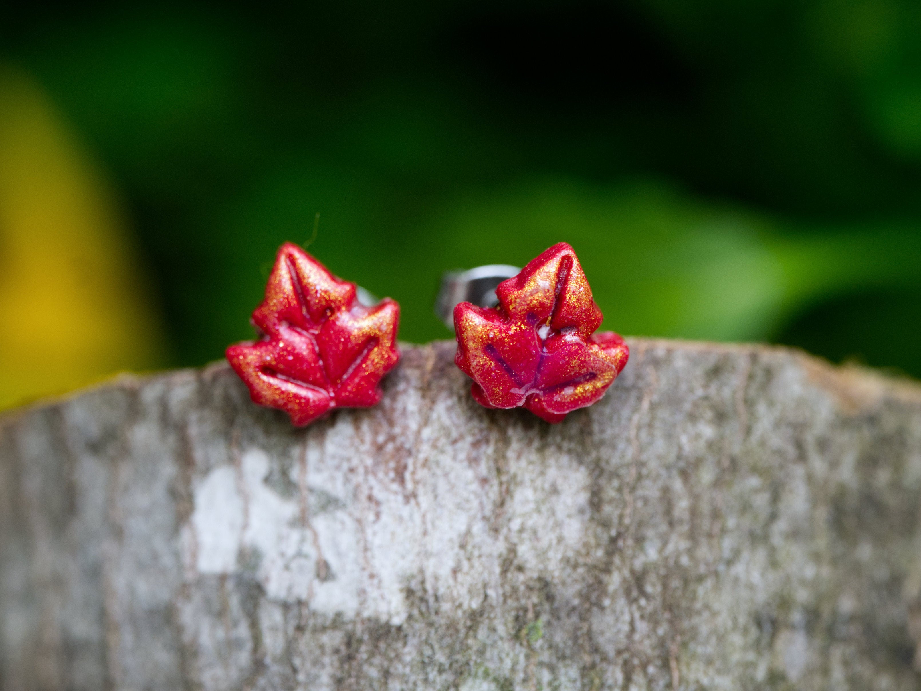 Close-up of red and gold shimmer maple leaf stud earrings perched on weathered tree bark with blurred green forest background