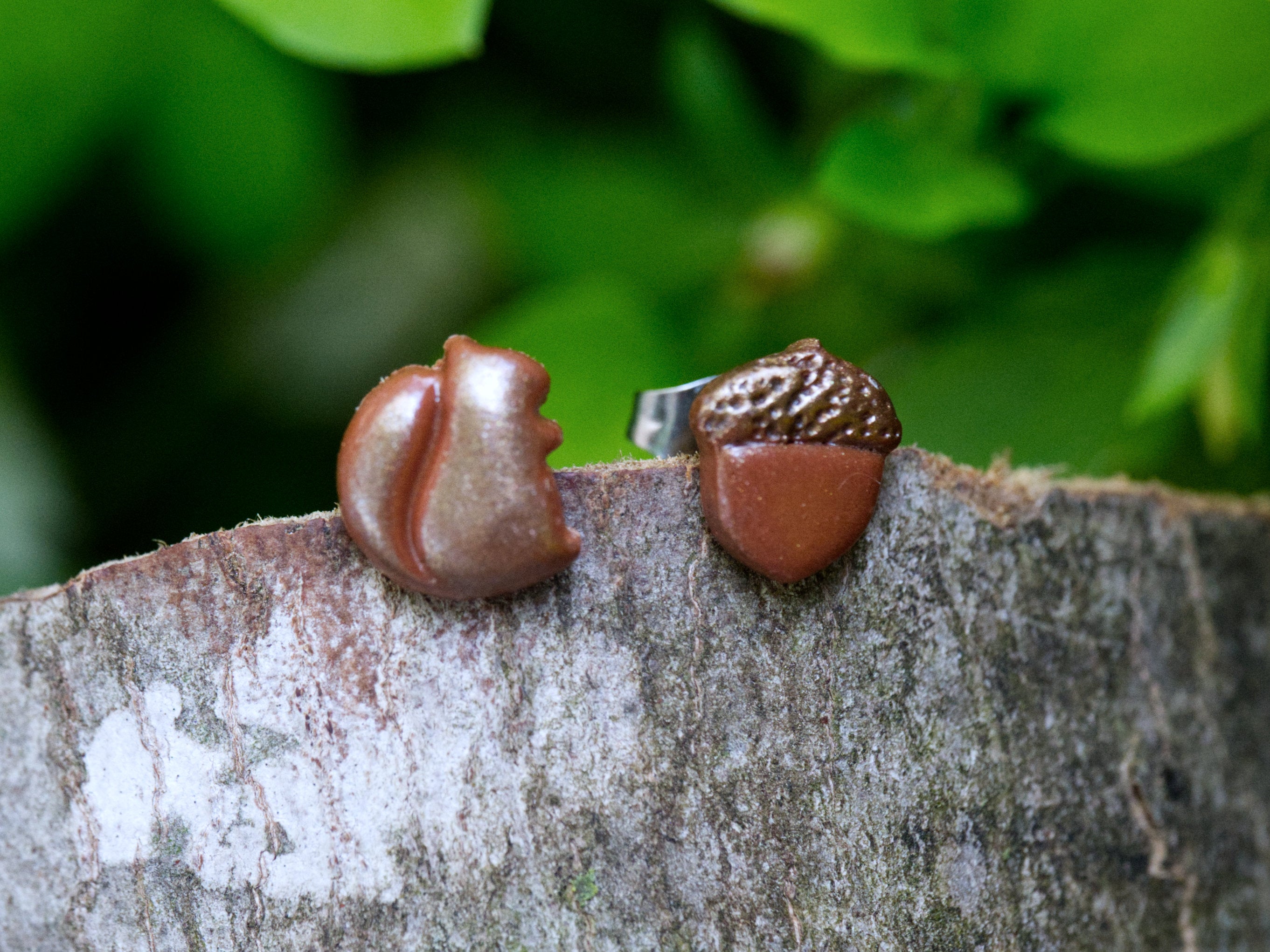Handmade mismatched brown polymer clay squirrel and acorn stud earrings perched on natural bark surface with green garden foliage background