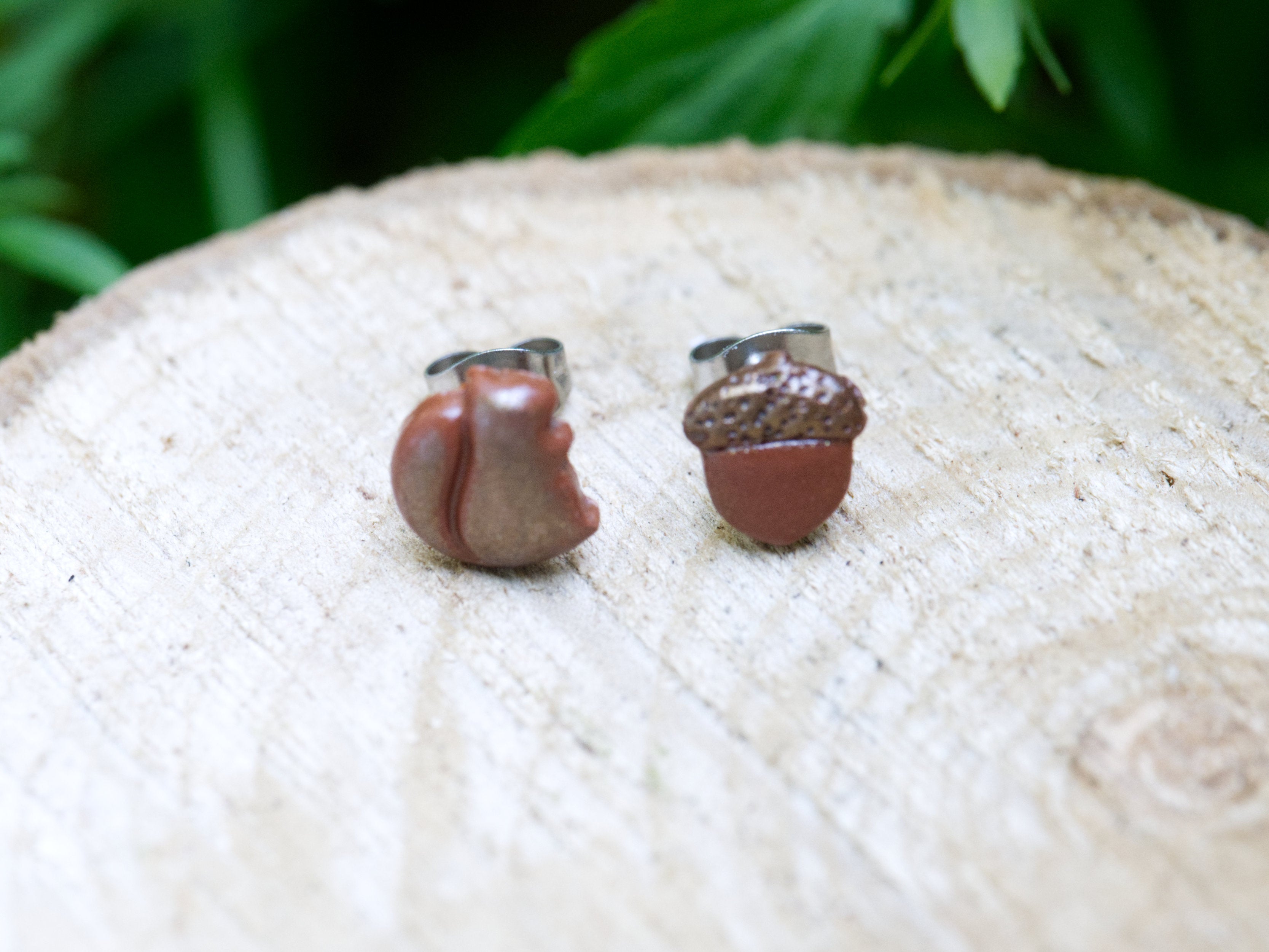 Close-up of mismatched squirrel and acorn polymer clay stud earrings showing sculpted squirrel features and textured acorn cap detail