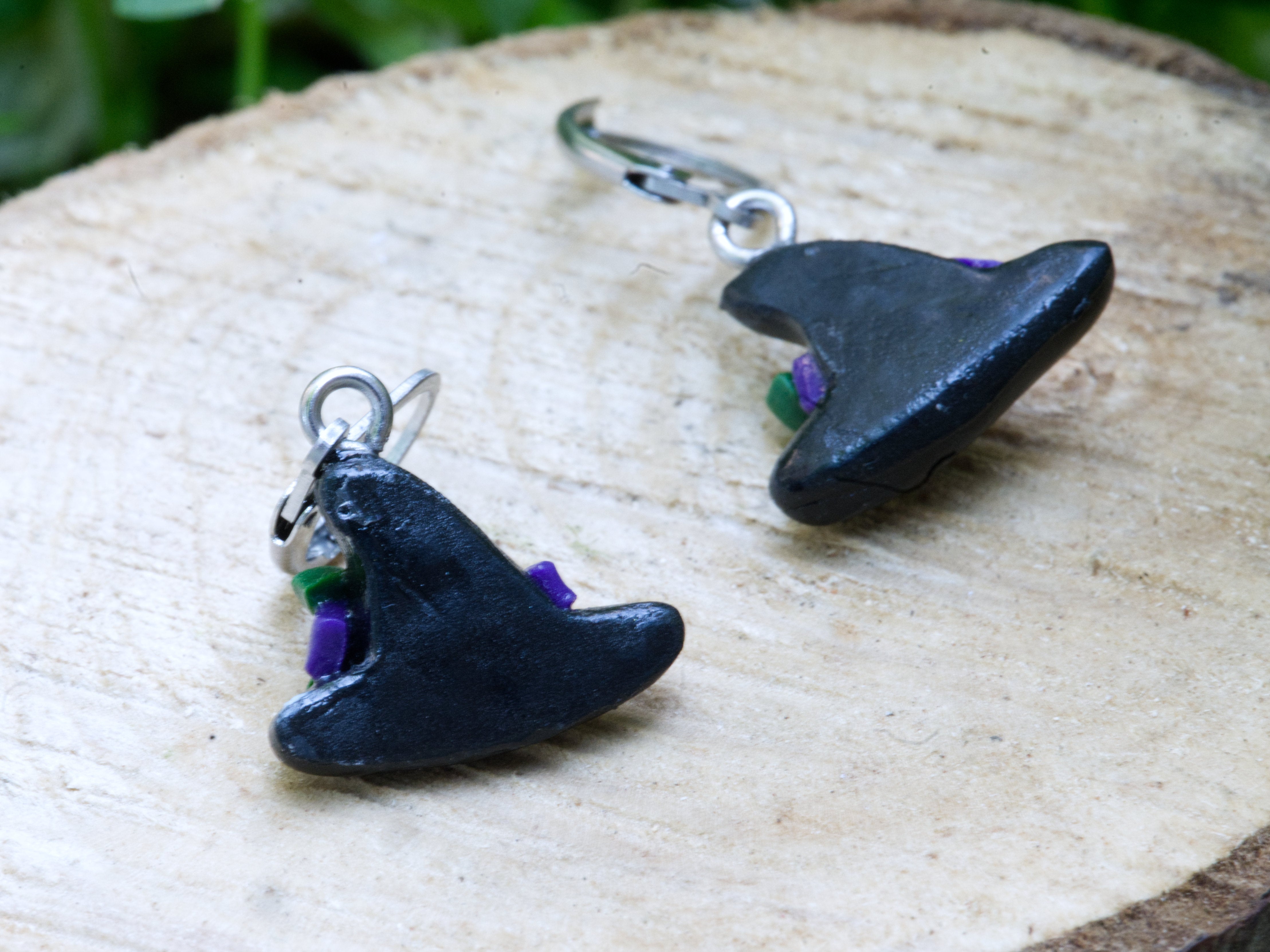 Back of witch hat polymer clay earrings on wood slice with soft green foliage and white flower bokeh background