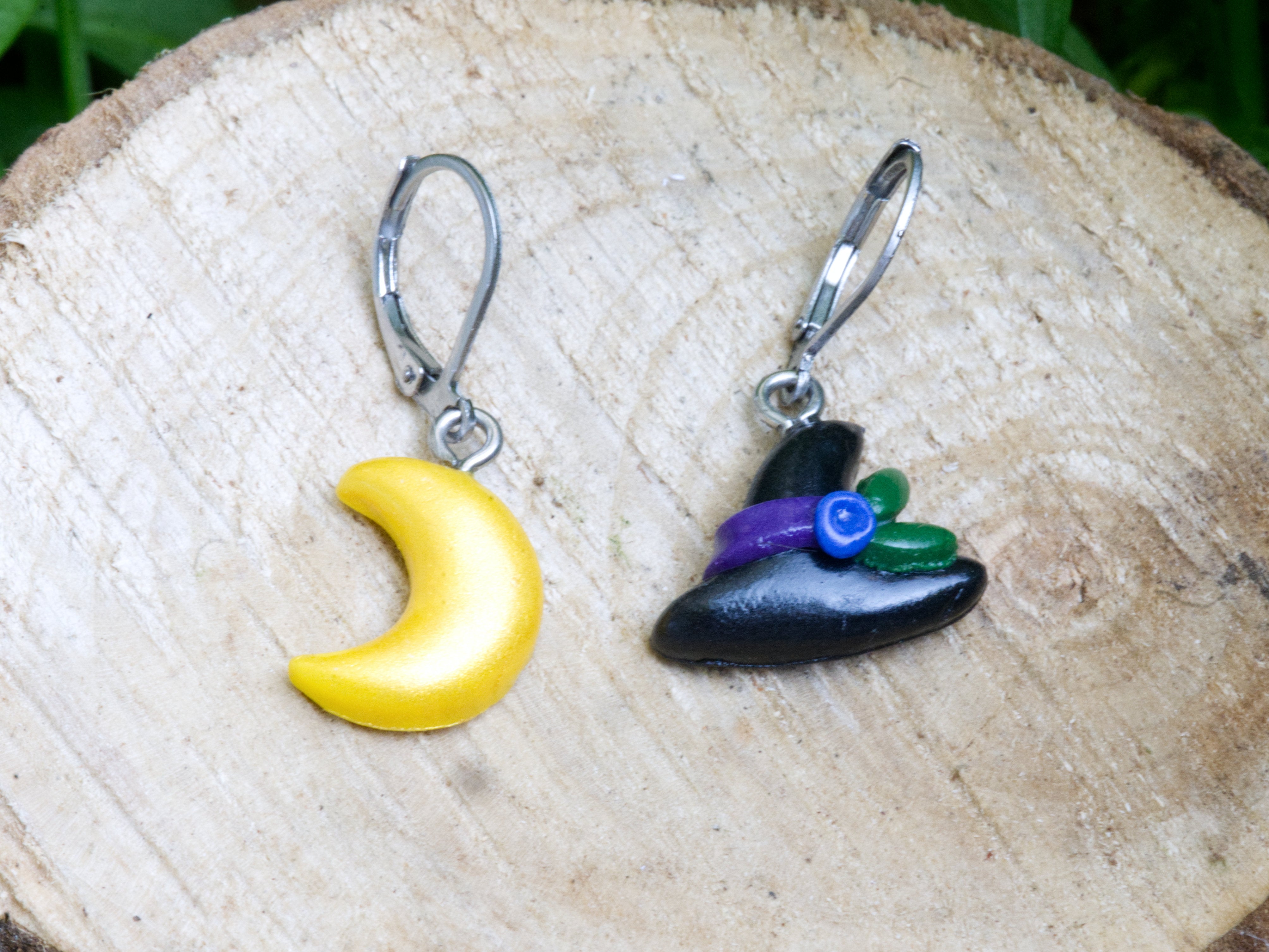 Handmade mismatched black polymer clay witch hat and gold shimmer crescent moon leverback earrings resting on natural wood slice with garden flowers