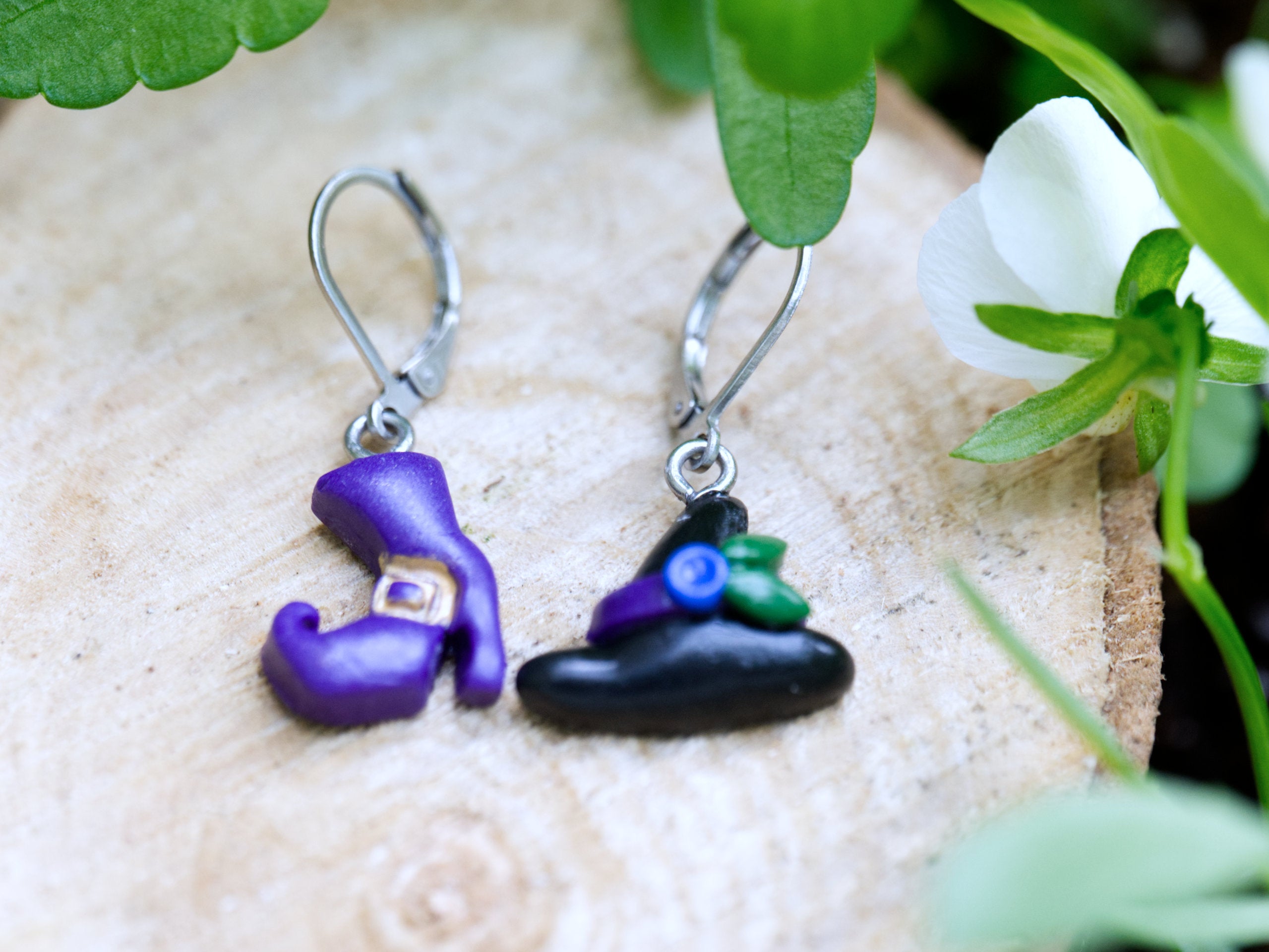 Handmade mismatched black polymer clay witch hat and purple boot leverback earrings resting on natural wood surface with green foliage background