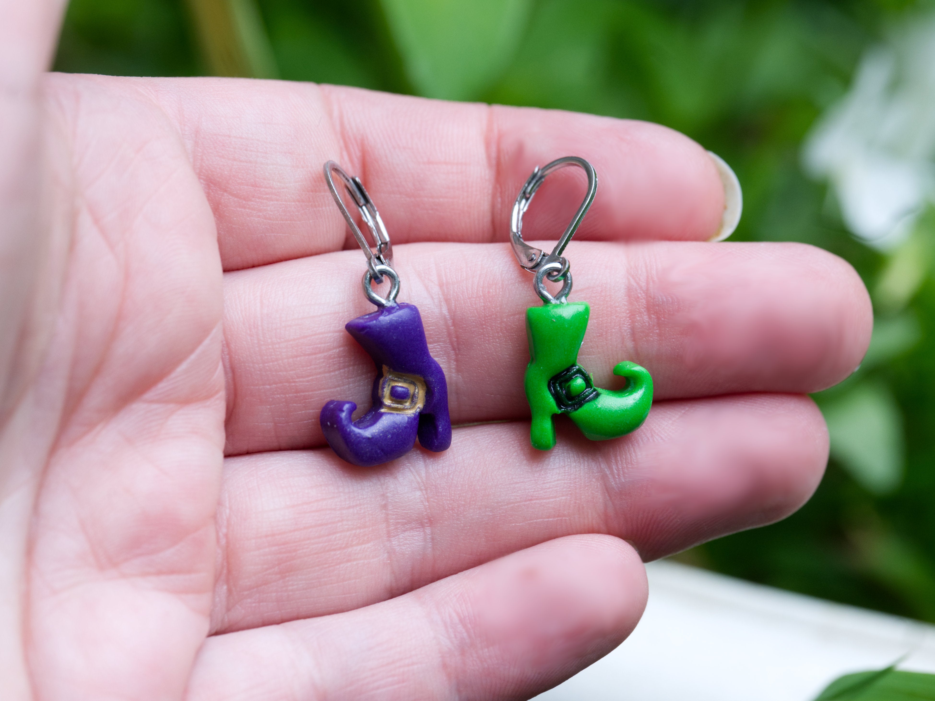 Mismatched purple and green polymer clay witch boot earrings on hand to demonstrate size