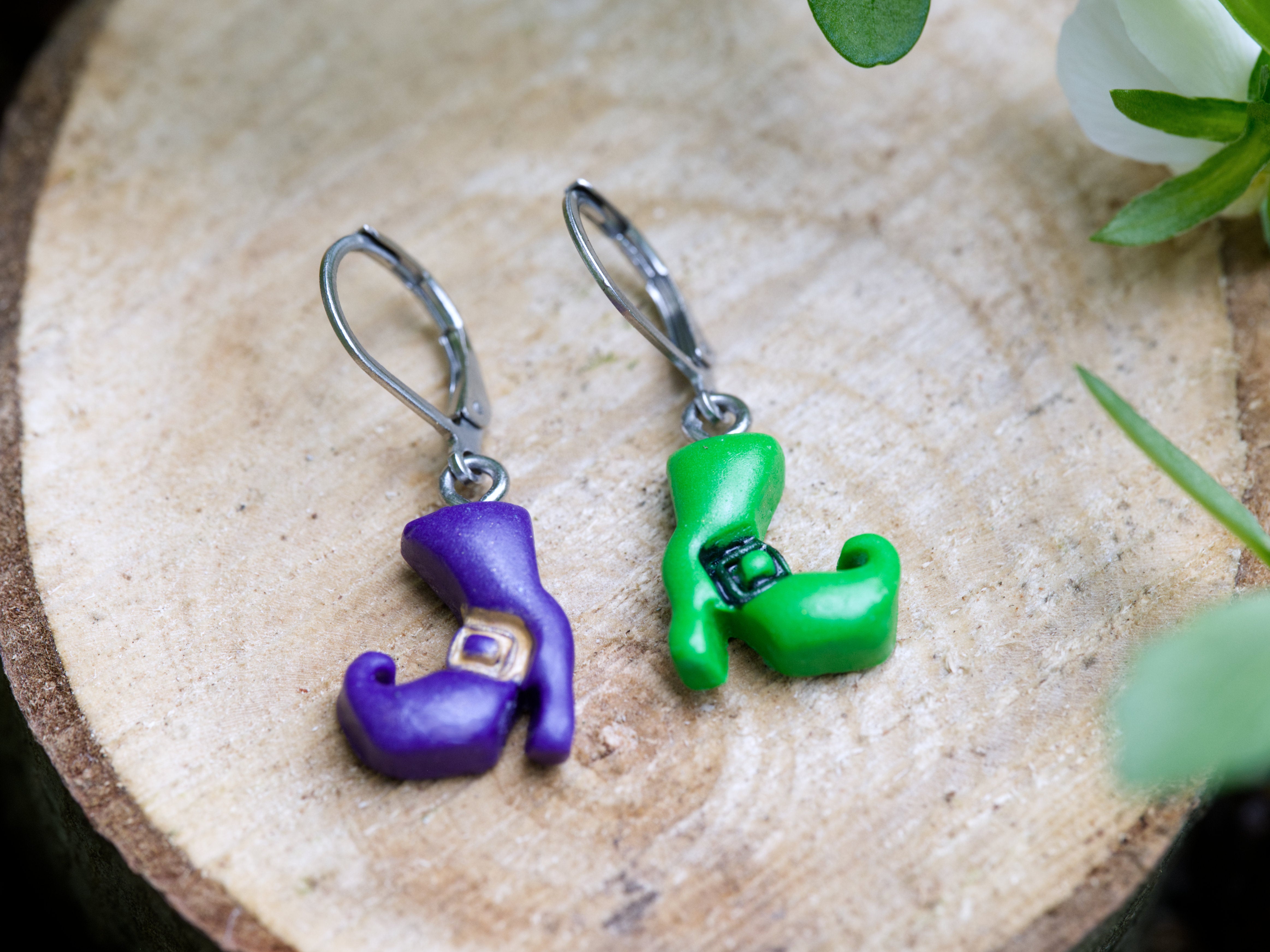 Handmade mismatched purple and green polymer clay witch boot earrings on silver leverback hooks resting side by side on natural wood slice with floral background