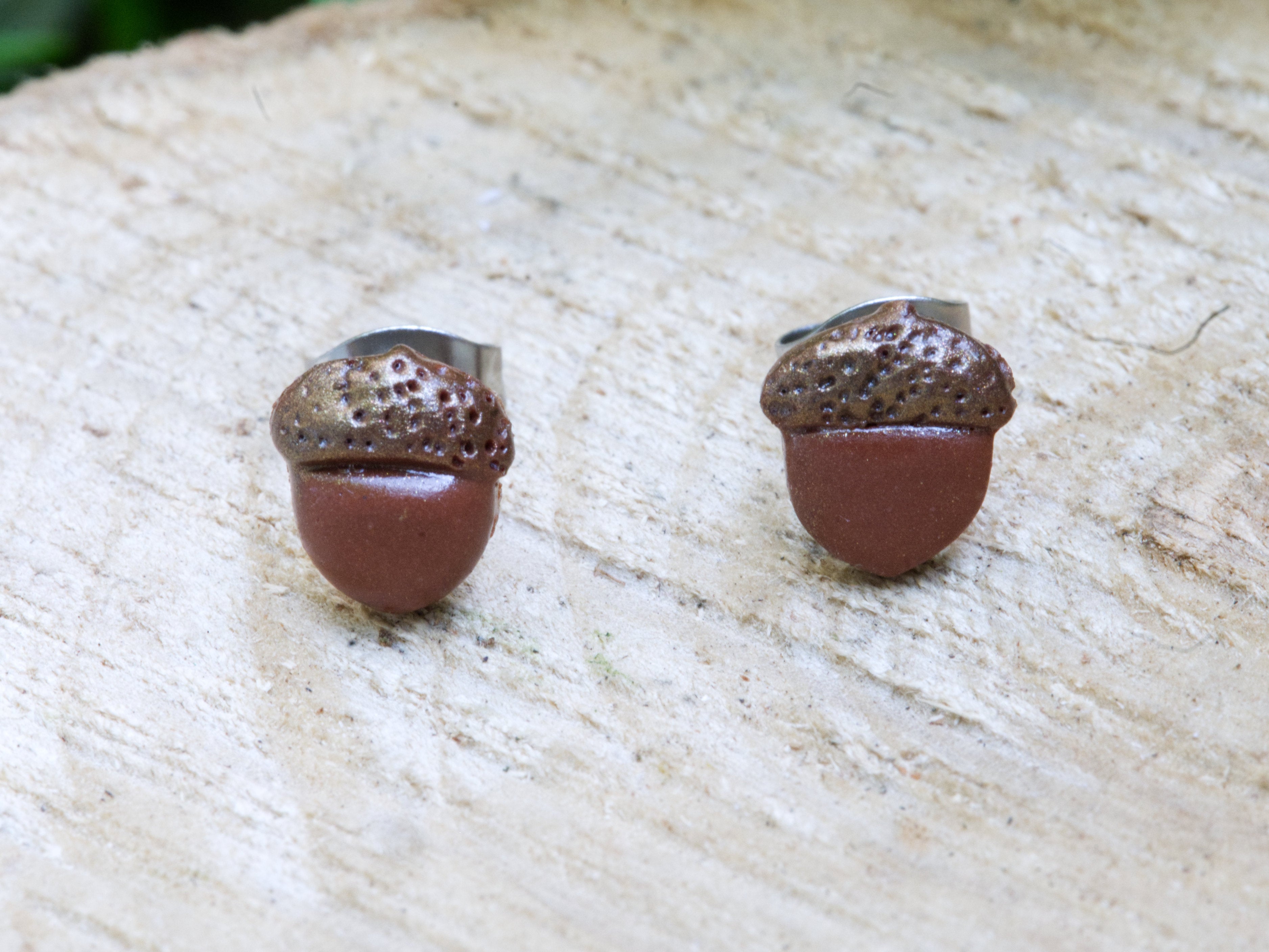 Handmade brown polymer clay acorn stud earrings perched on natural wooden surface with green garden foliage background