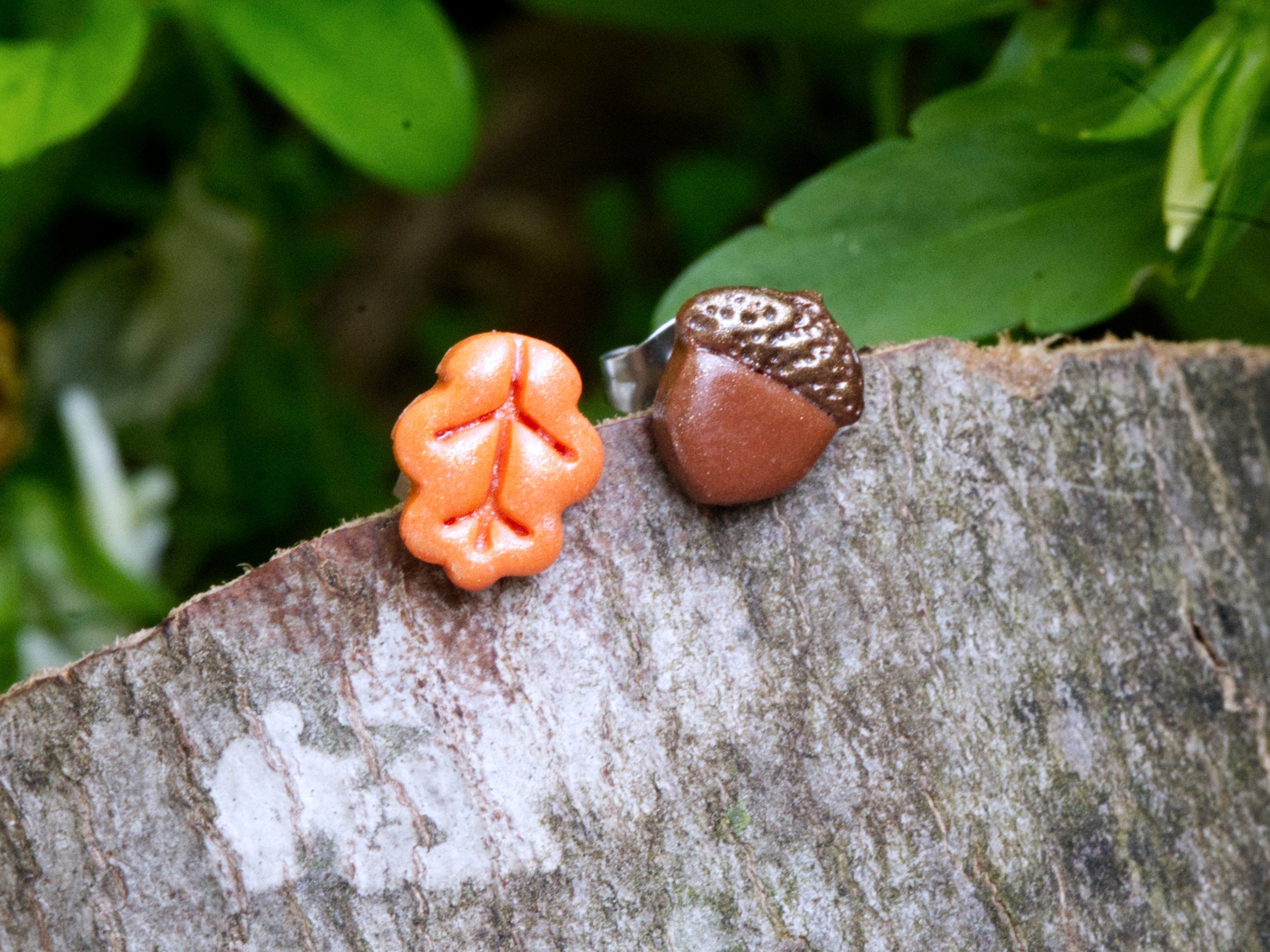 Handmade mismatched orange polymer clay oak leaf and brown acorn stud earrings perched on natural bark surface with green garden foliage background