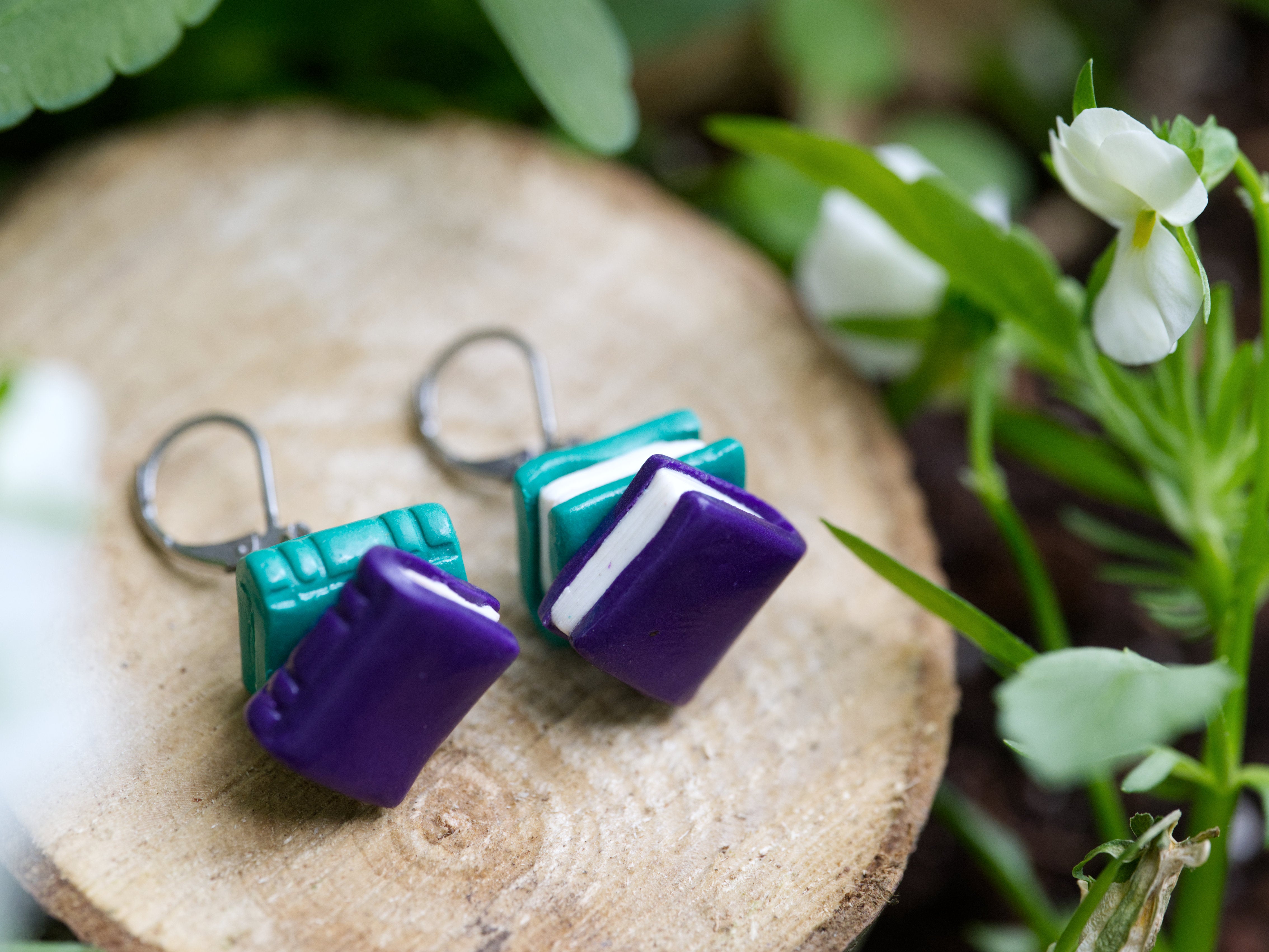Teal and purple book stack earrings resting on natural wood slice surrounded by green leaves and white flowers in soft natural daylight