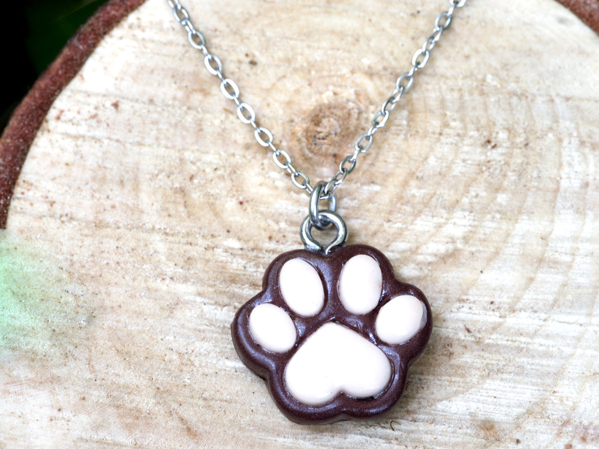 Brown paw print necklace with a handmade coffee-brown polymer clay pendant, pink pads, and stainless steel chain displayed on natural wood.
