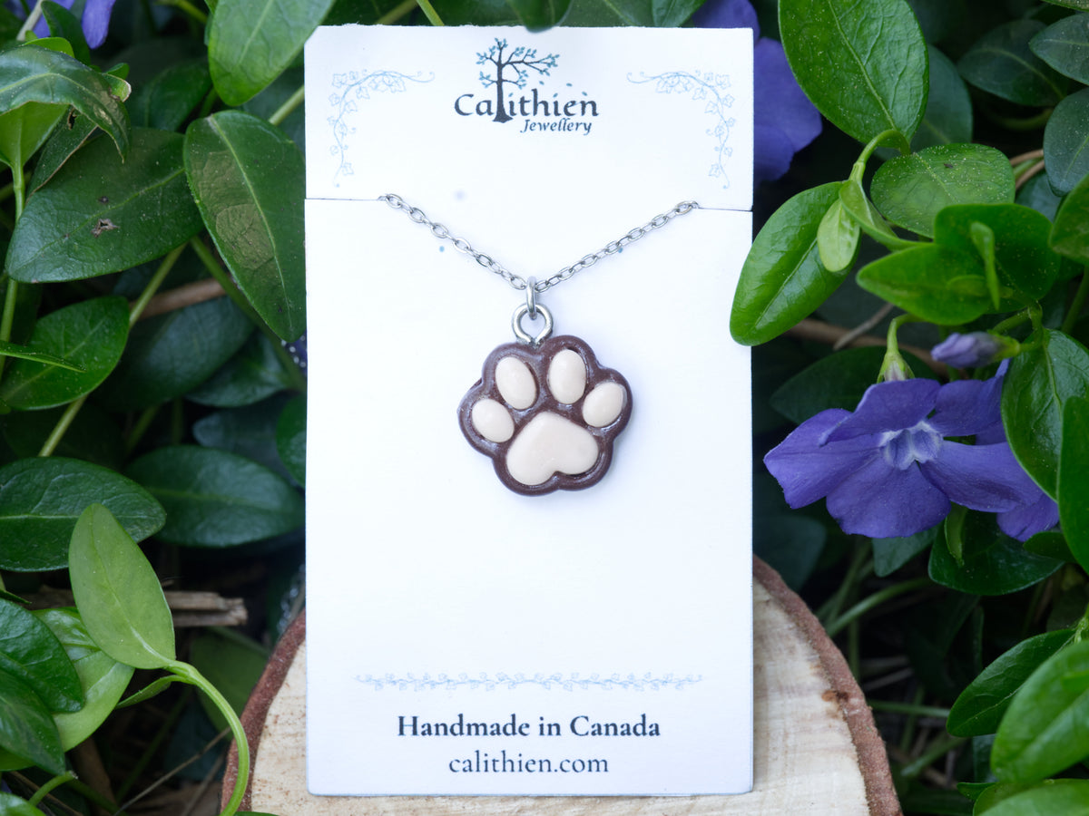 Brown paw print necklace displayed on its branded jewellery card against a floral backdrop.