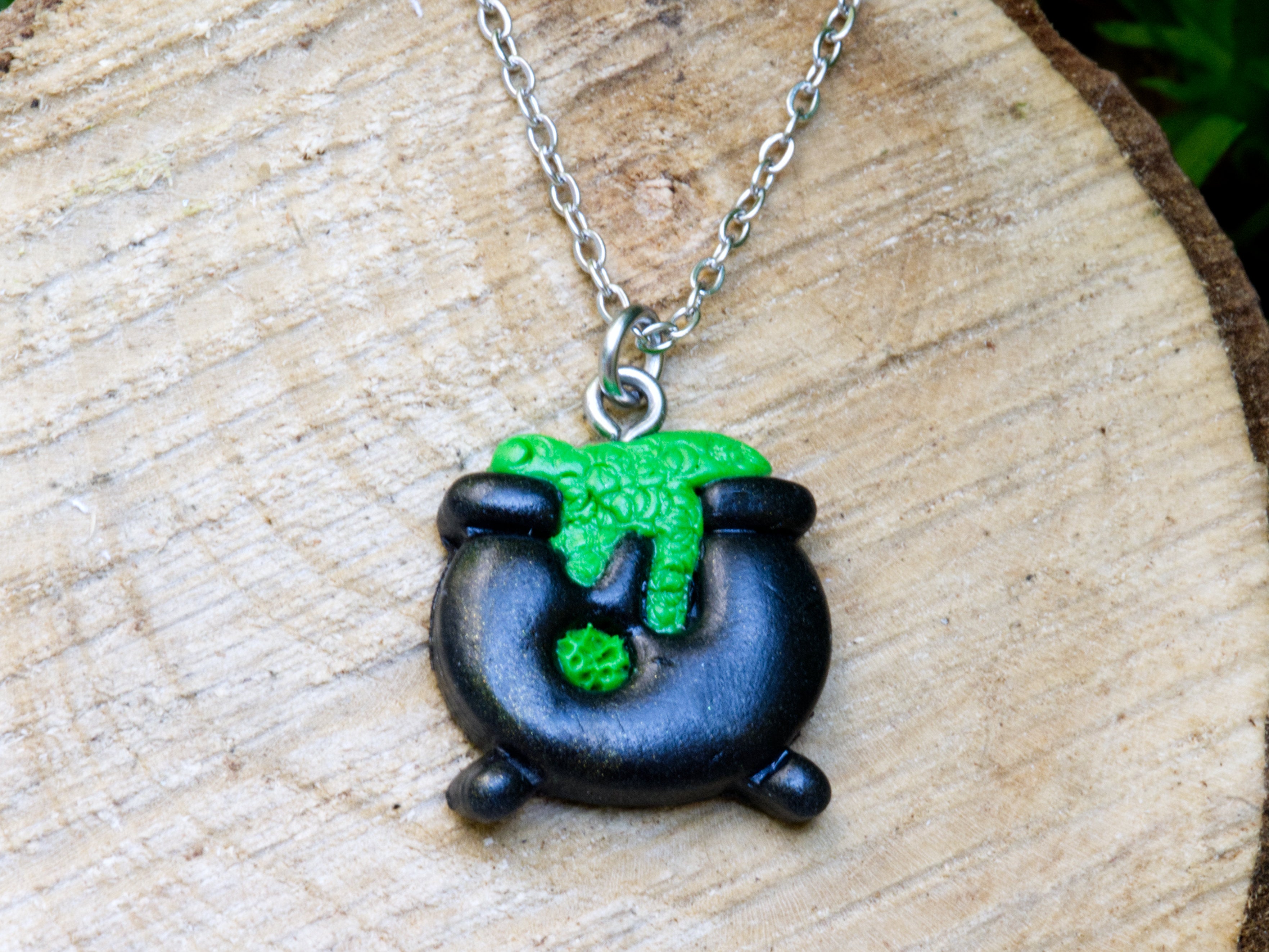 Handmade black polymer clay cauldron pendant necklace with bubbling green brew detail on 304 stainless steel chain on natural wood surface