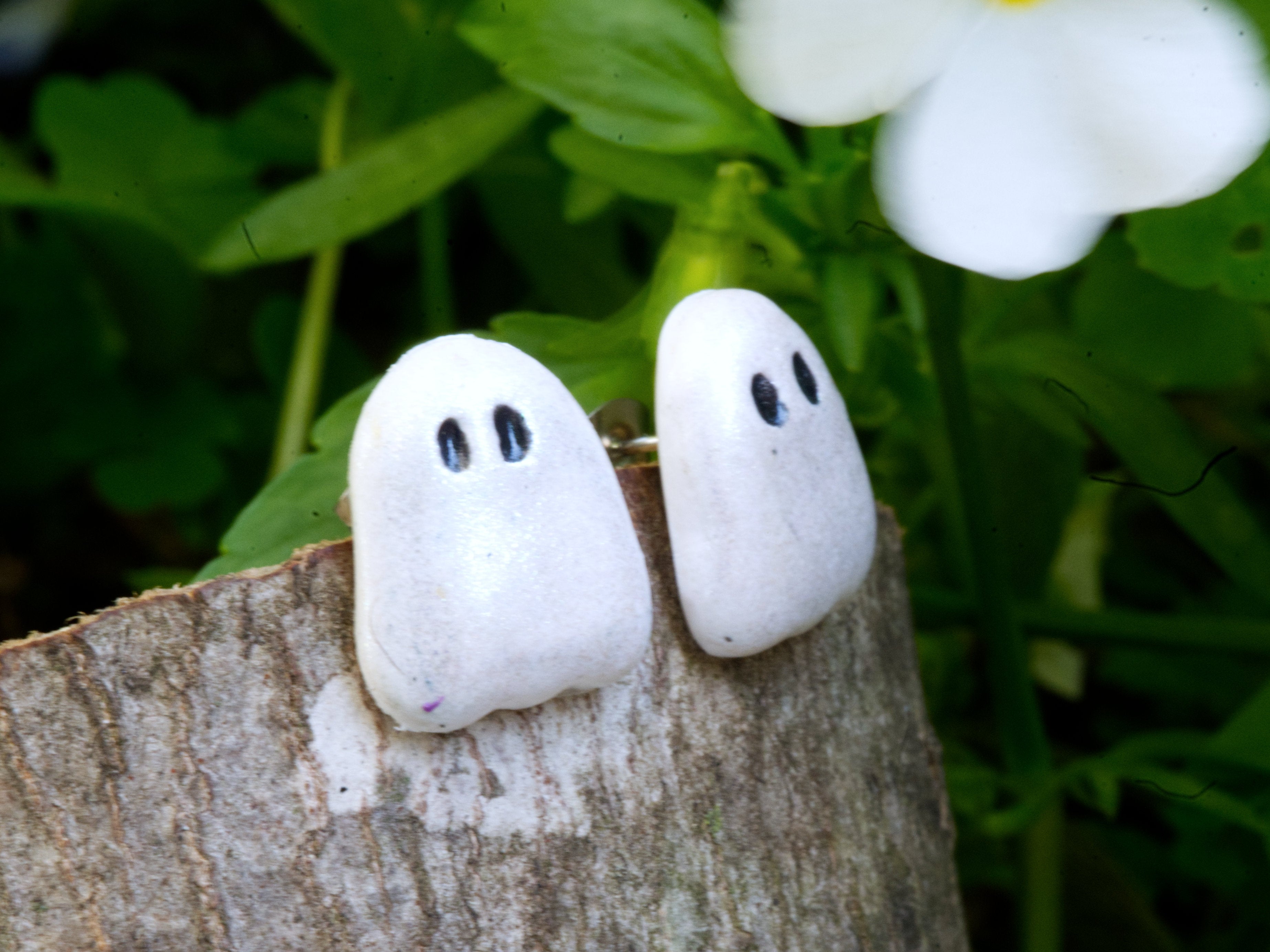 Pair of ghost stud earrings on wood slice surface with soft green bokeh background in natural daylight