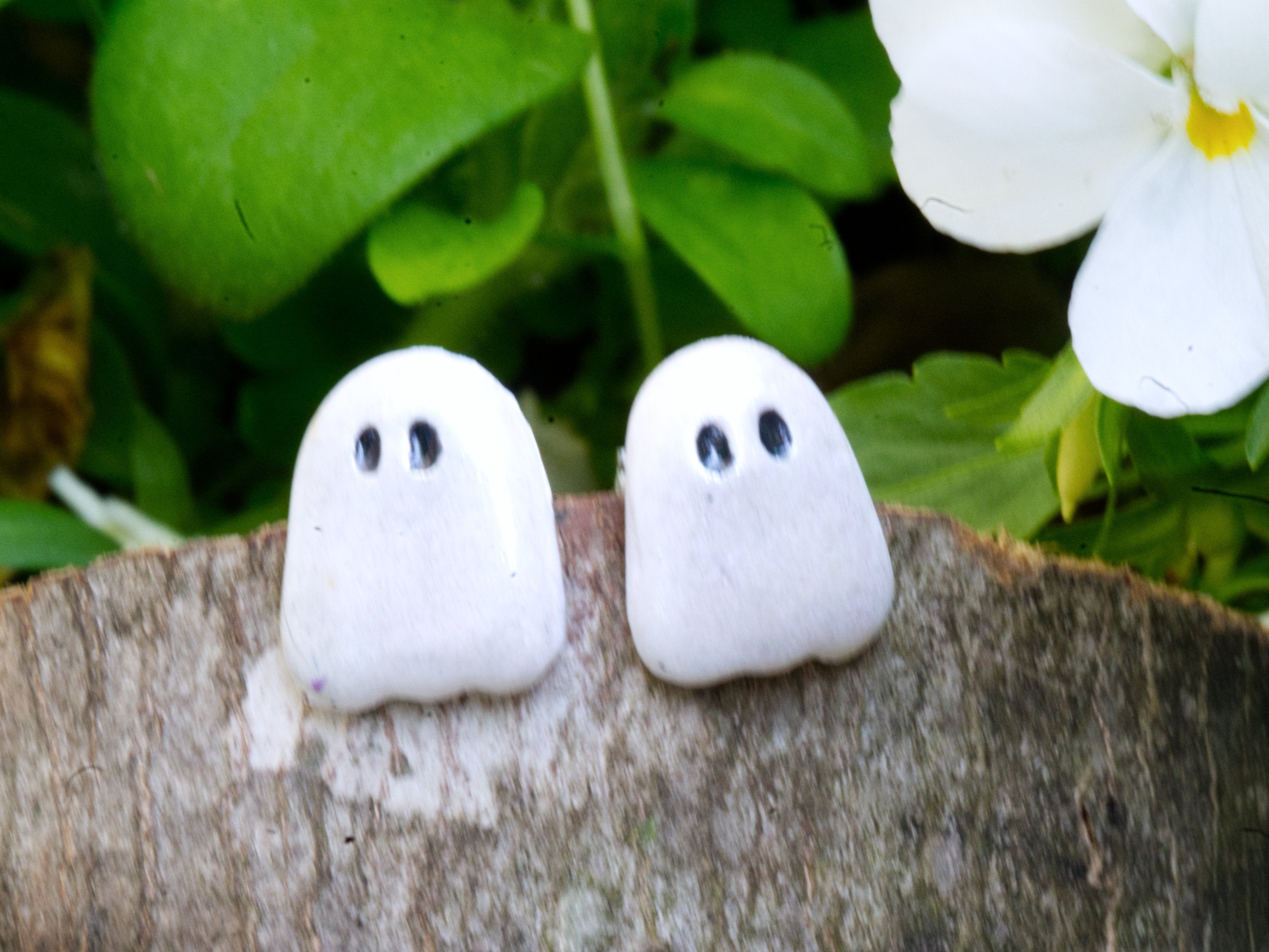 Handmade white pearl shimmer polymer clay ghost stud earrings sitting side by side on natural wood slice with green garden foliage background