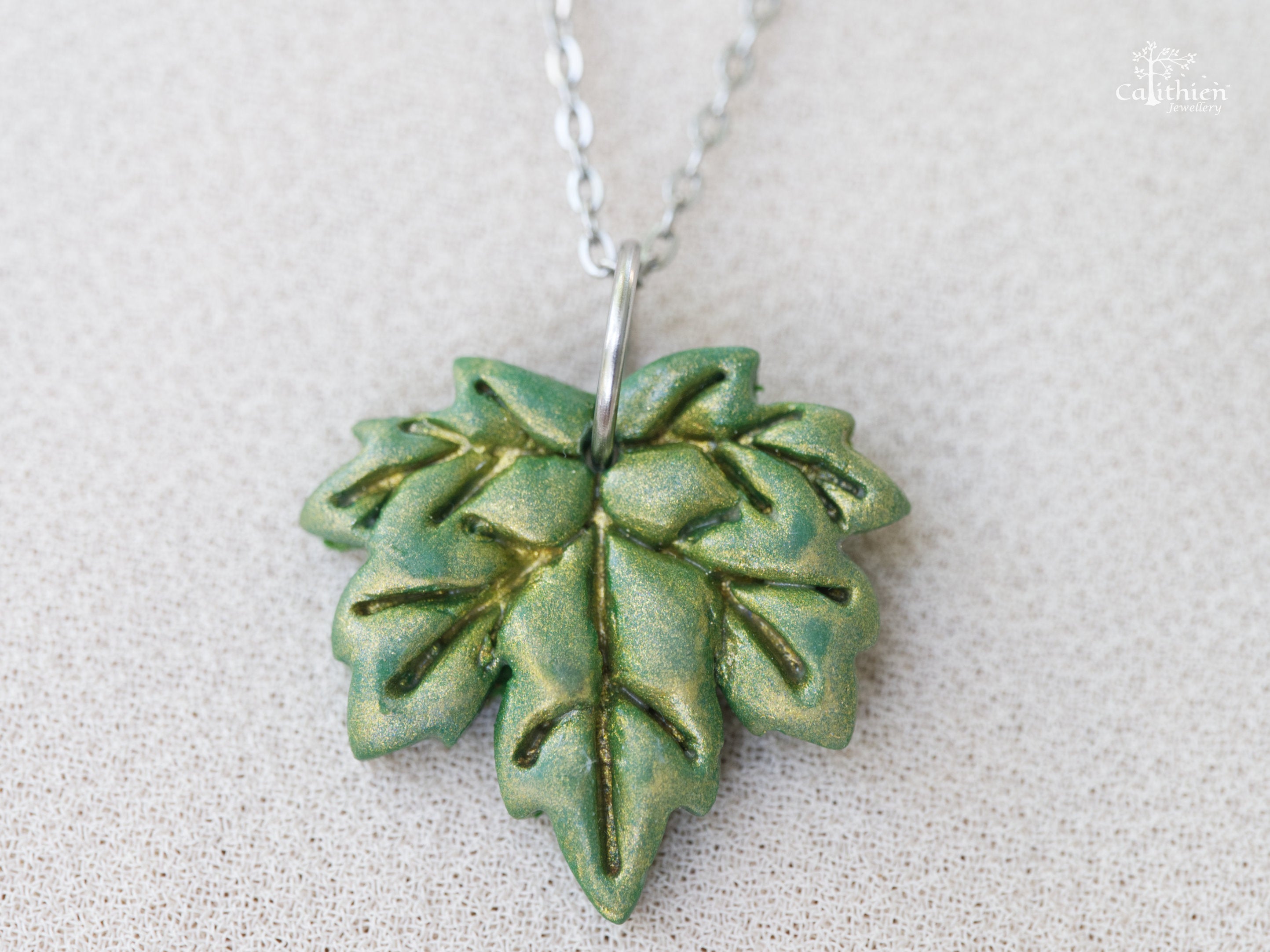 Handmade green shimmer polymer clay maple leaf pendant necklace on 304 stainless steel chain resting on white fabric
