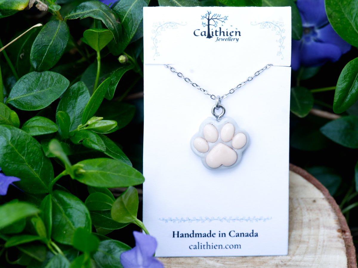 Grey paw print necklace displayed on its branded jewellery card against a floral backdrop.