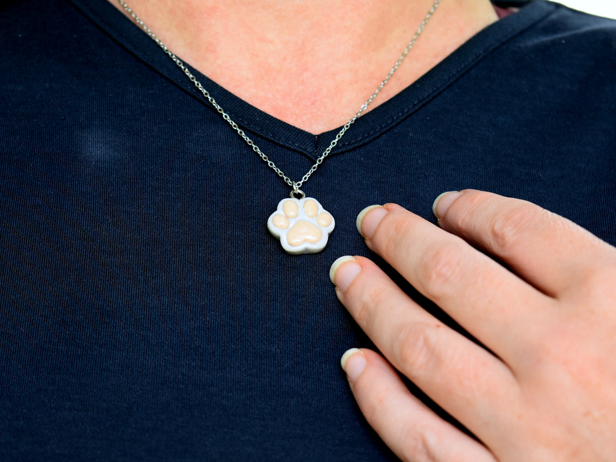 Handmade dove-grey polymer clay paw print necklace worn against a navy blue T-shirt, showing the pendant's size and everyday style.