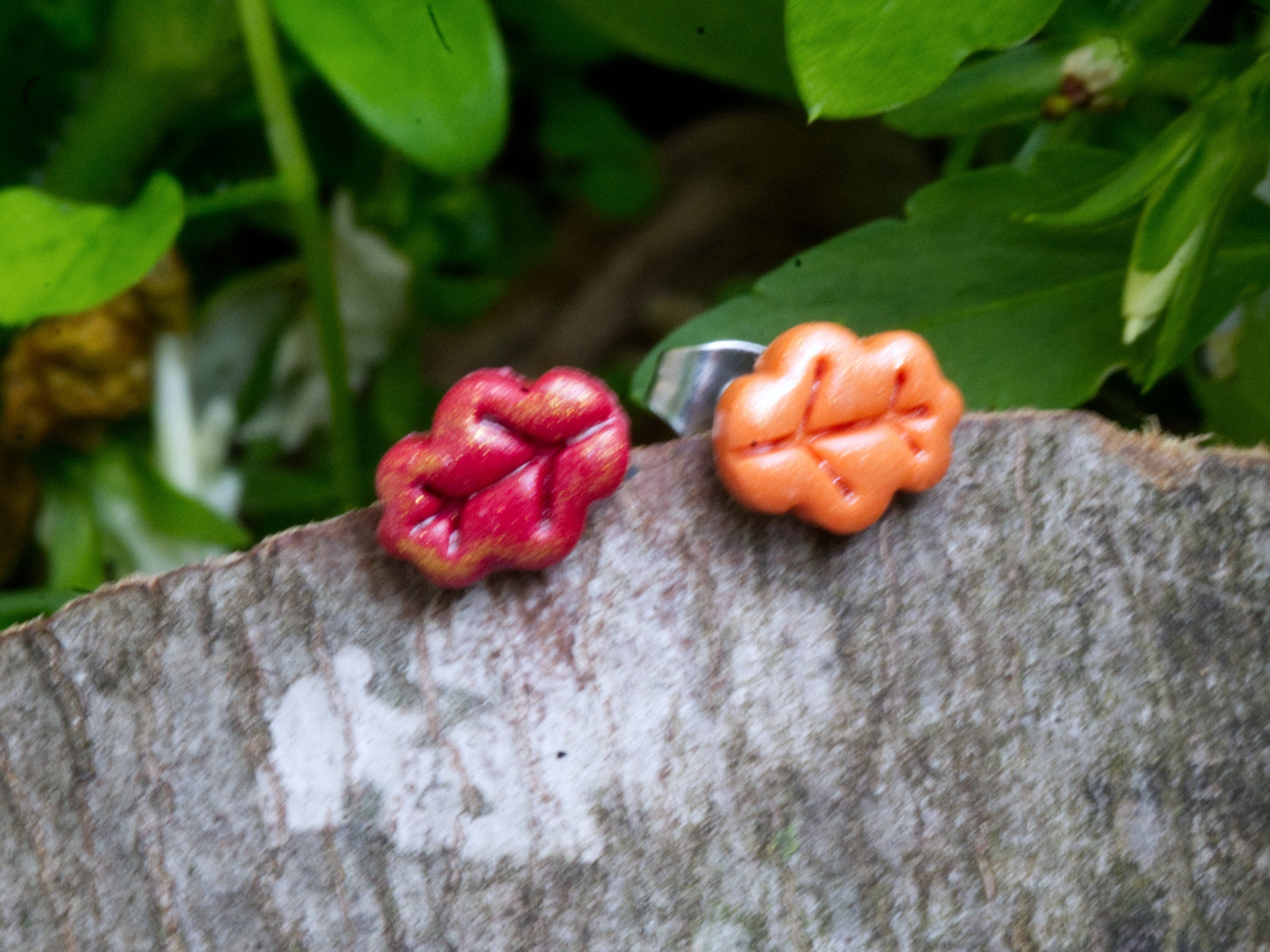 Handmade mismatched orange and red polymer clay oak leaf stud earrings perched on natural wood bark with green garden foliage background