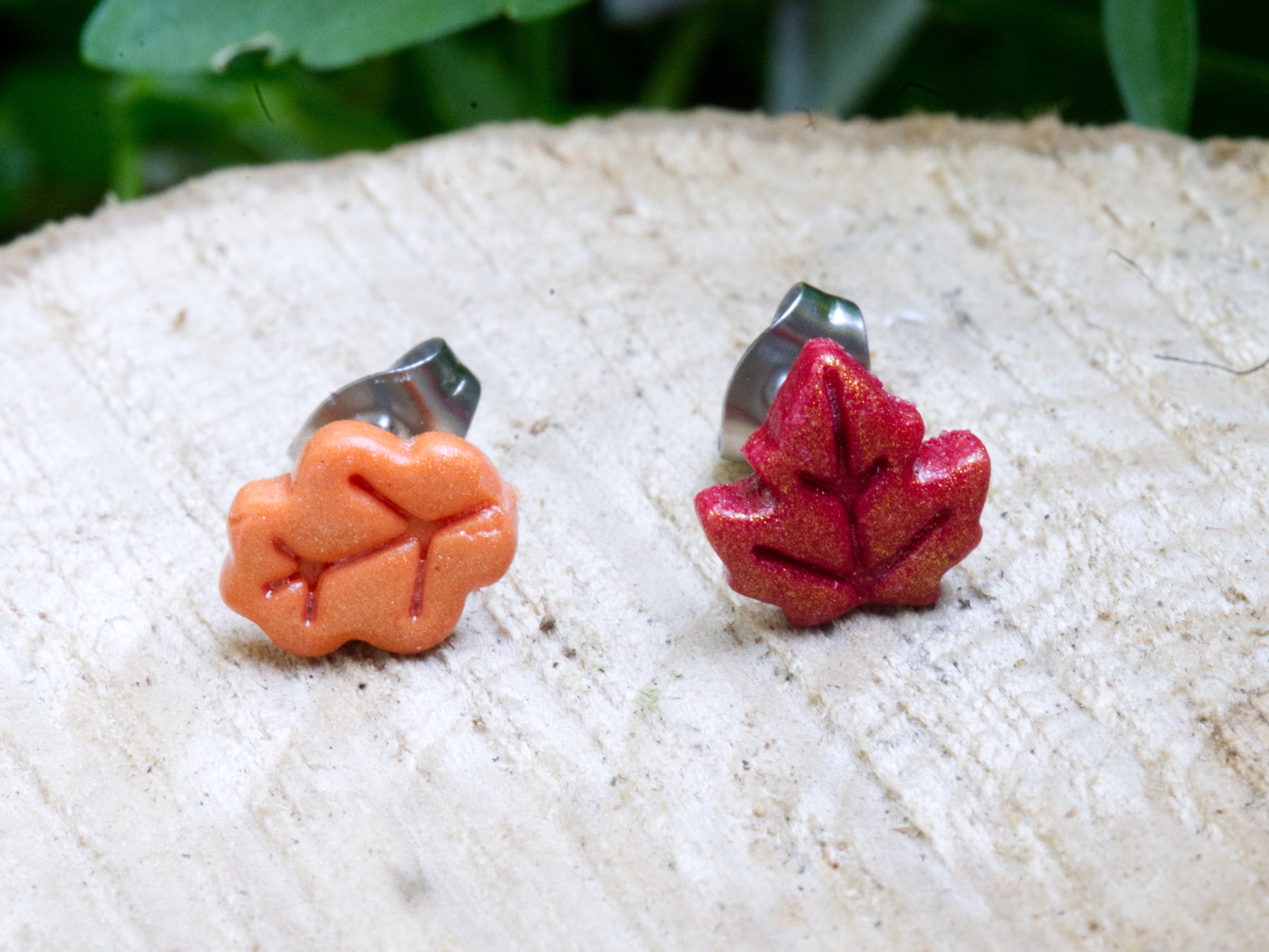Handmade mismatched orange oak leaf and red maple leaf polymer clay stud earrings on natural wood surface with green garden background