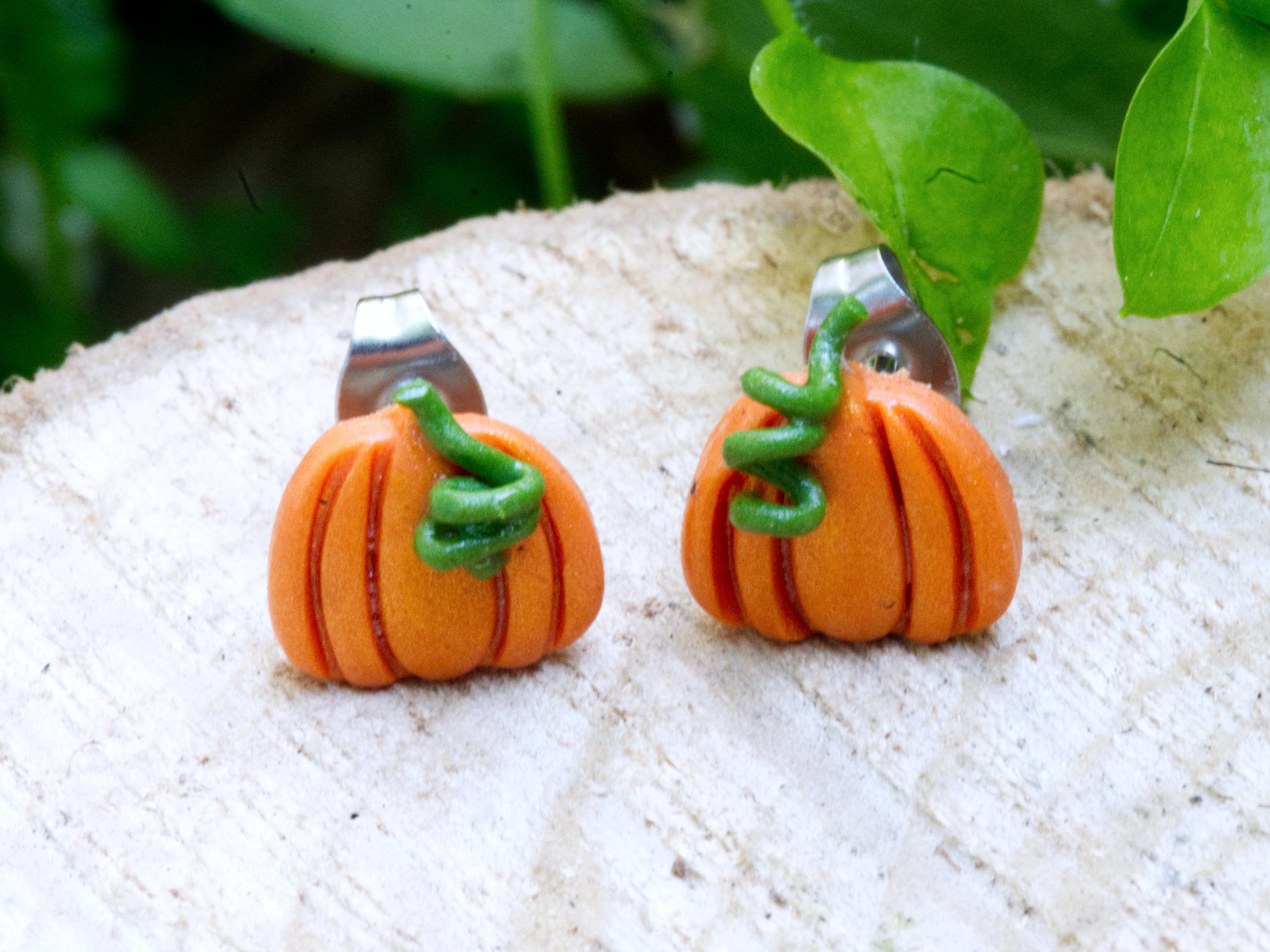 Pair of orange pumpkin stud earrings on wooden surface with soft green foliage bokeh and garden background