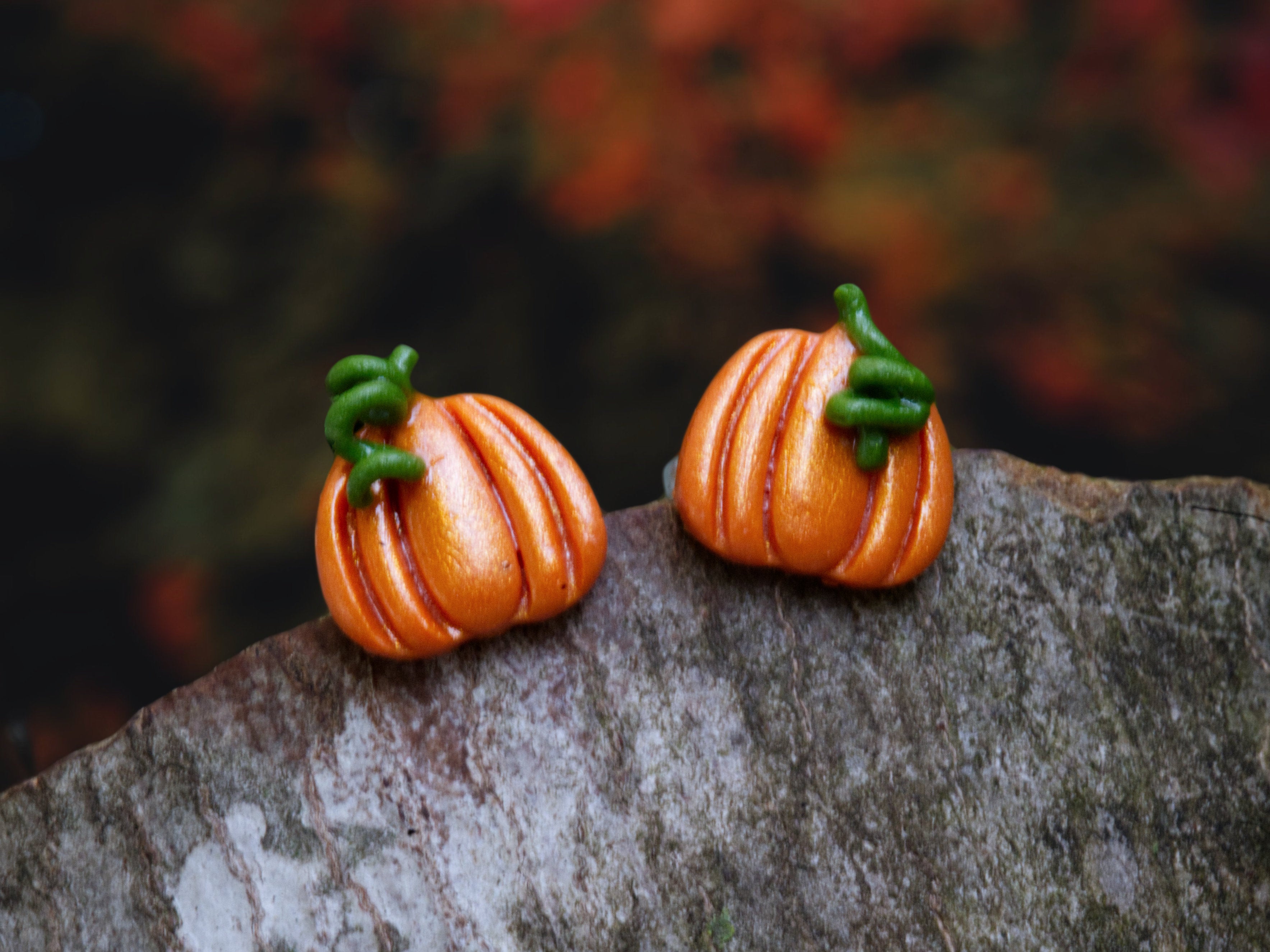 Handmade polymer clay pumpkin stud earrings in orange with shimmer and green stems, resting on natural bark with colourful autumn leaves in the background.