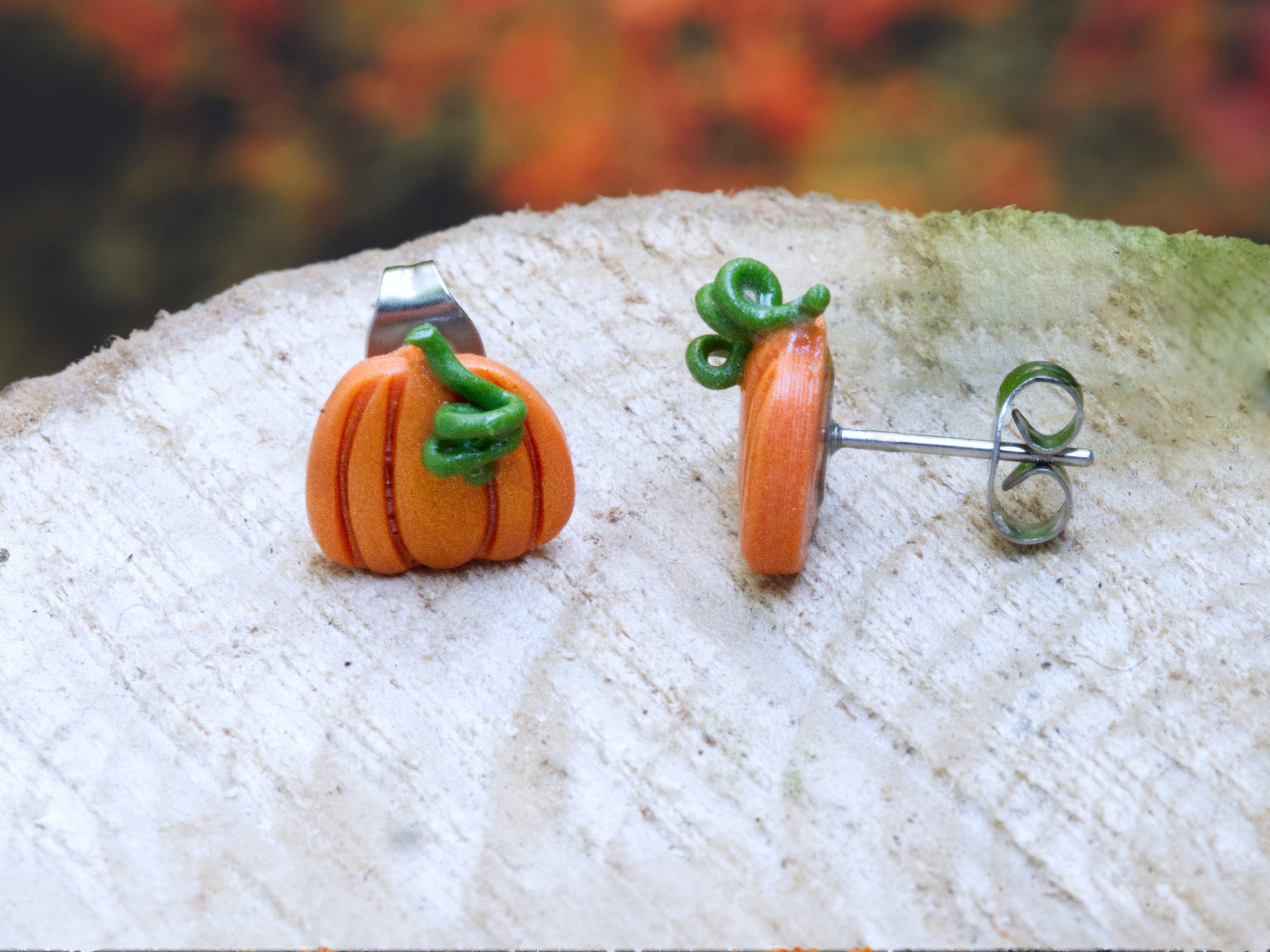 Handmade orange polymer clay pumpkin stud earrings with shimmering finish and green curly stems, shown from front and side views against a natural autumn-inspired background.