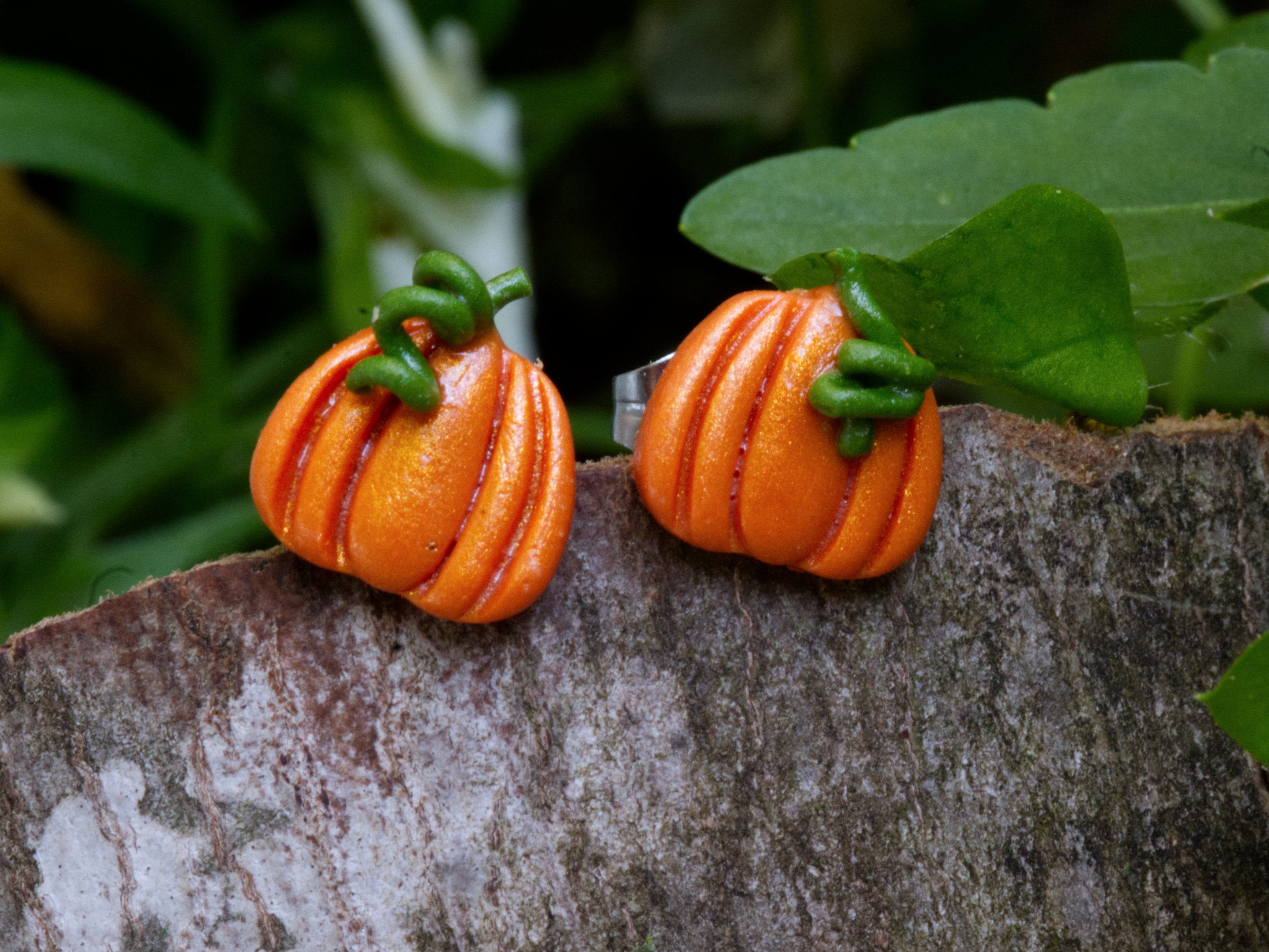 Handmade orange polymer clay pumpkin stud earrings with green stem and leaf detail perched on natural bark surface with garden foliage background