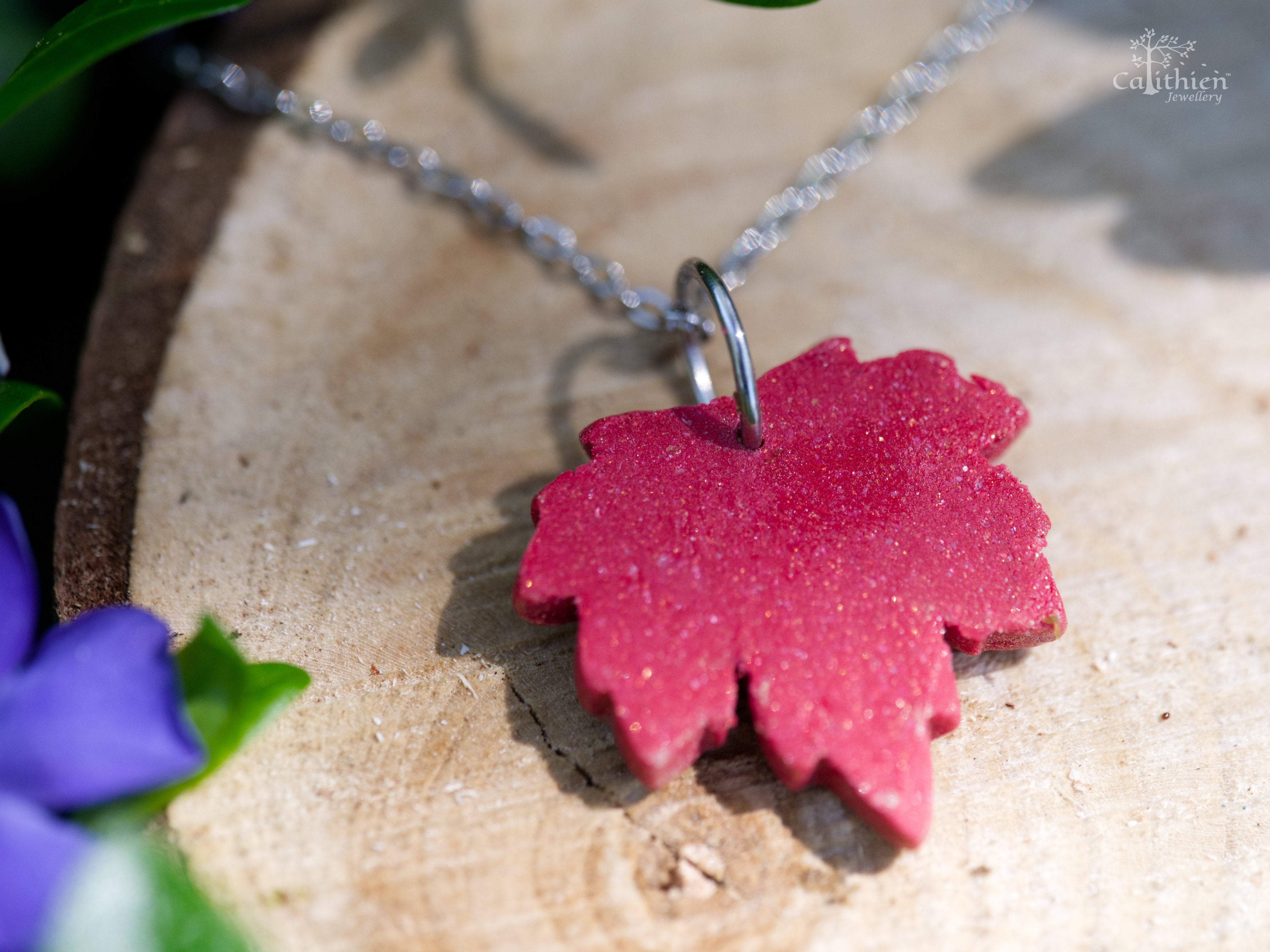 Reverse view of handmade red and gold shimmer polymer clay maple leaf pendant necklace on 304 stainless steel chain resting on natural wood slice with green foliage