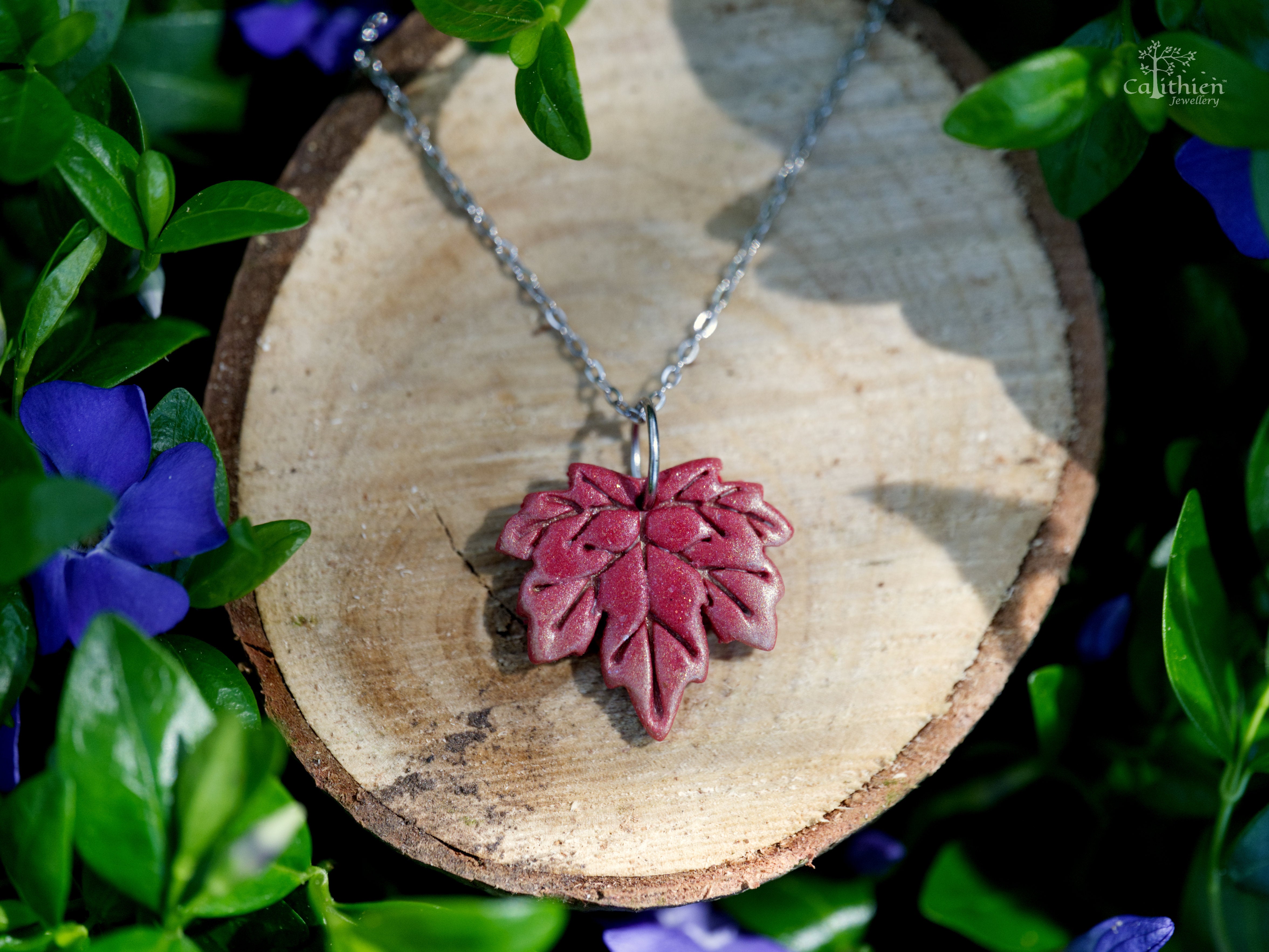 Red maple leaf necklace pendant on wood surface surrounded by garden greenery with soft light bokeh background