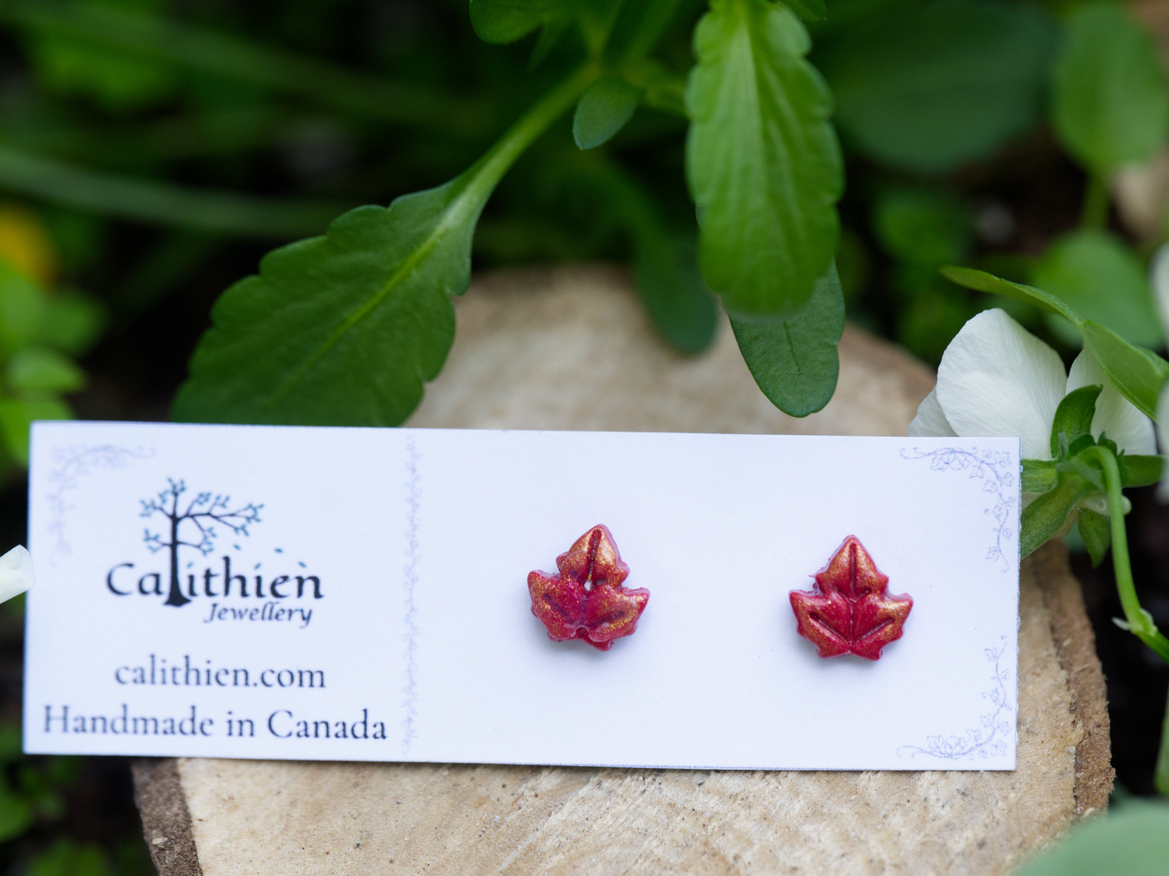Red polymer clay maple leaf stud earrings on Calithien Jewellery branded display card propped on stone with white garden flowers